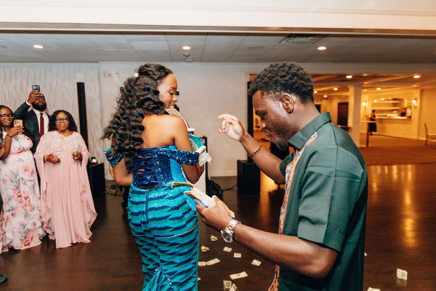 Guest in teal outfit spraying money on a woman in a blue dress on the wedding dance floor.