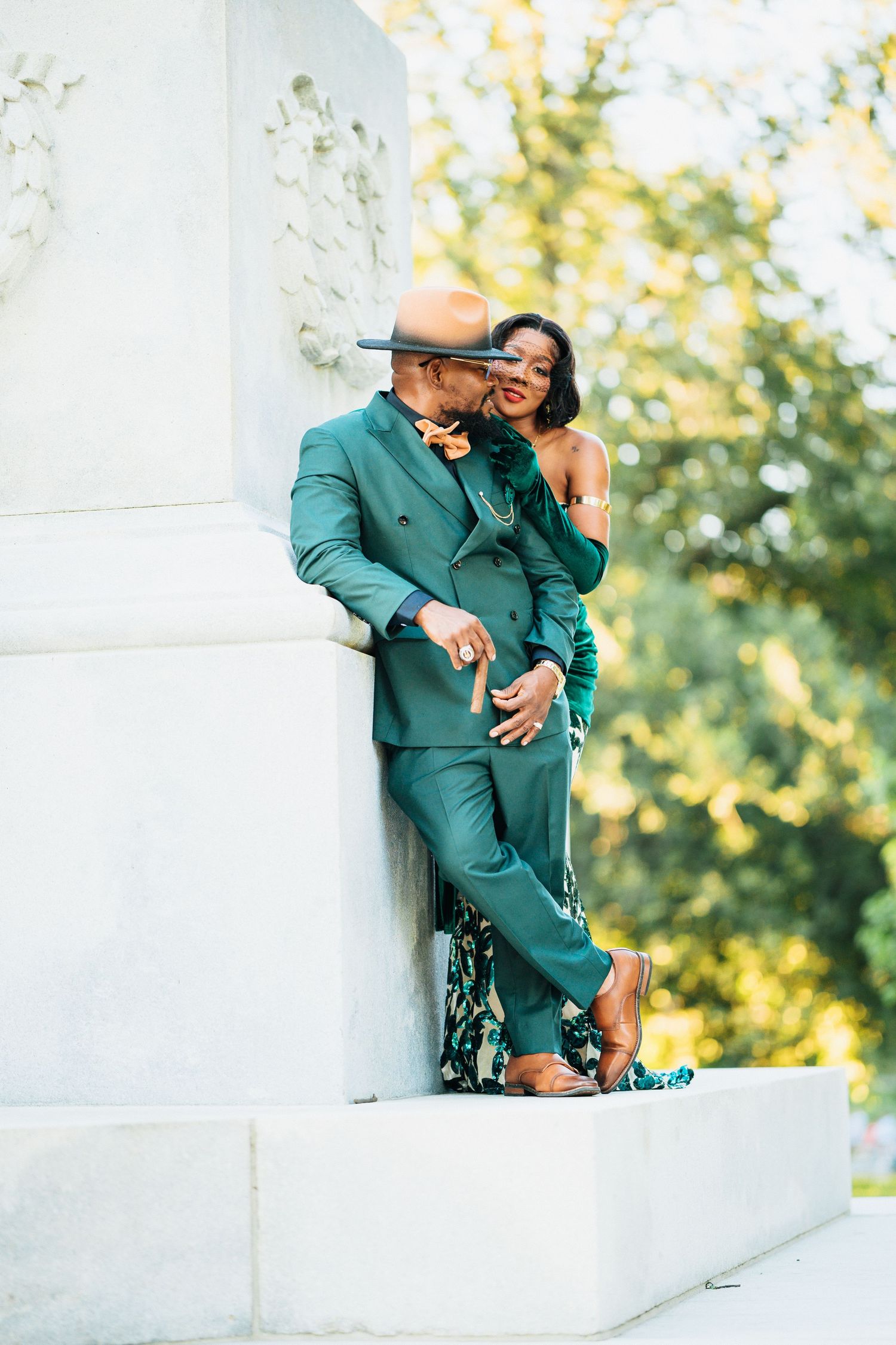 Elegant Black couple embracing by white stone pillar in green park setting.