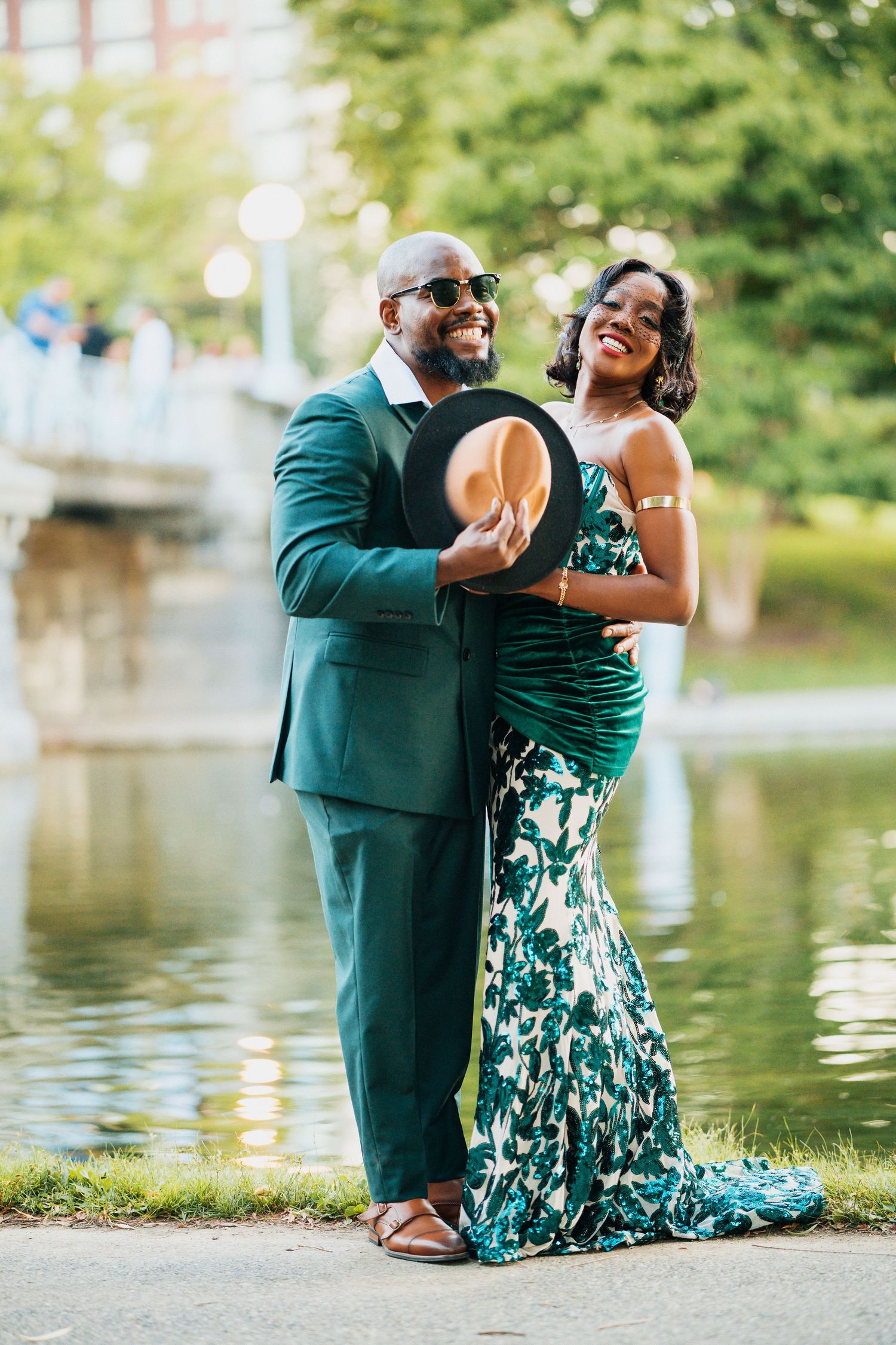Smiling couple in green formal wear posing by reflective pond in park.
