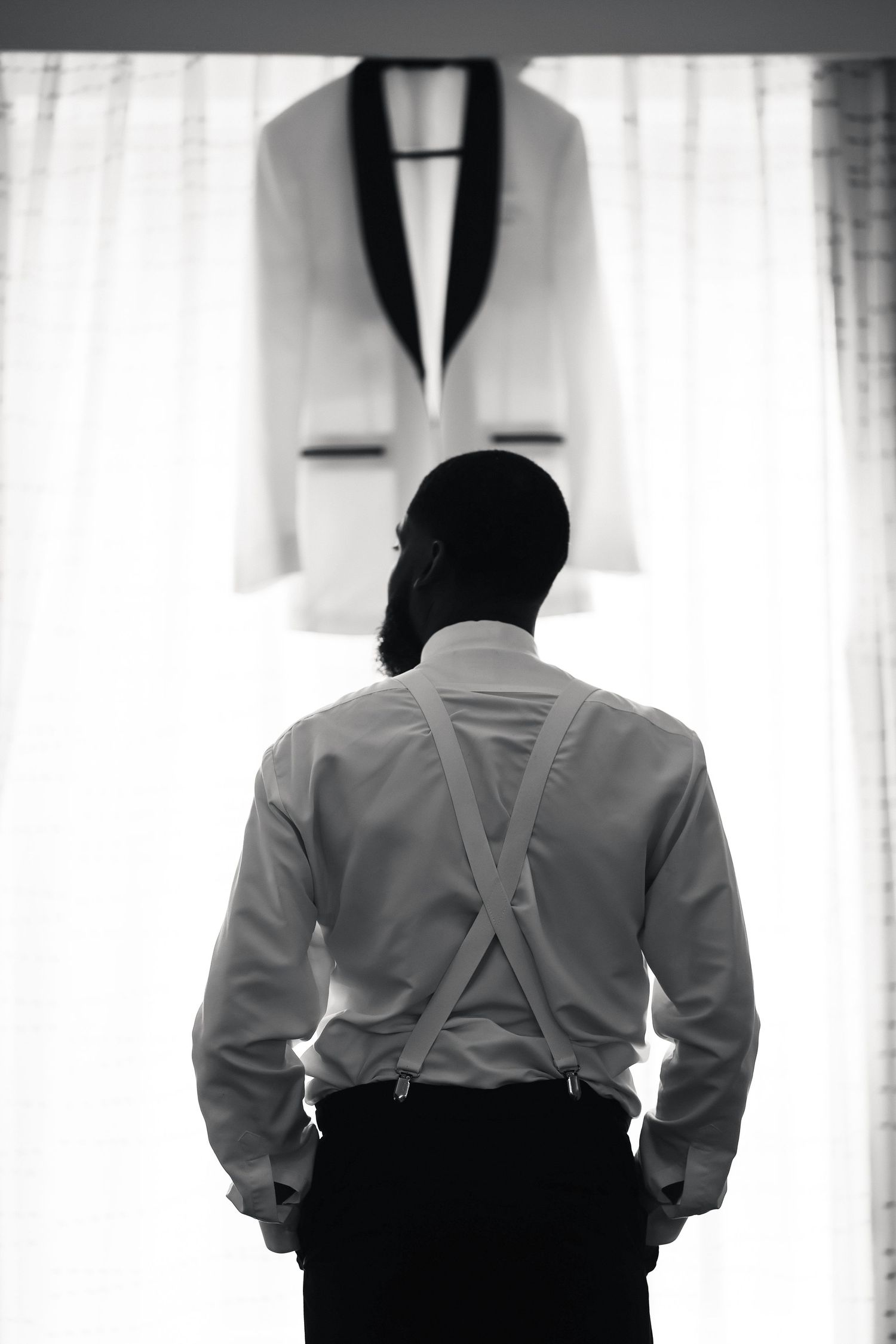 Black and white photo of a groom in suspenders looking at his white tuxedo jacket hanging by a window.