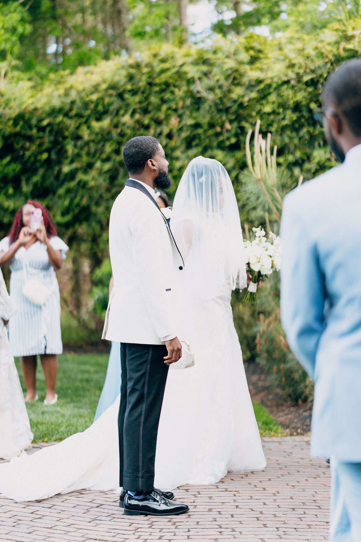 Groom in white tuxedo stands with bride in white gown and veil during outdoor wedding ceremony.
