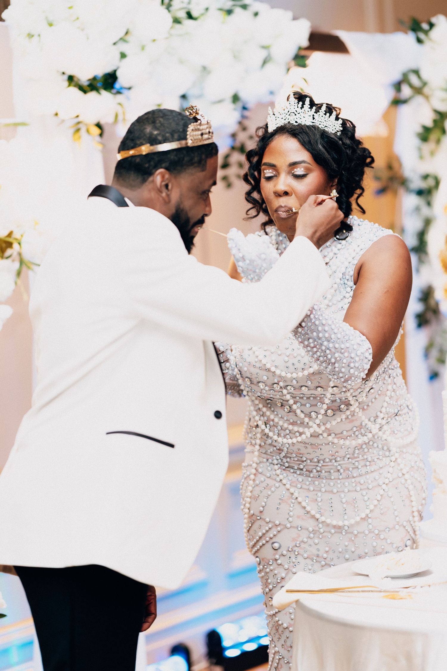 Groom in white tuxedo feeding wedding cake to bride wearing pearl gown and tiara at elegant reception.