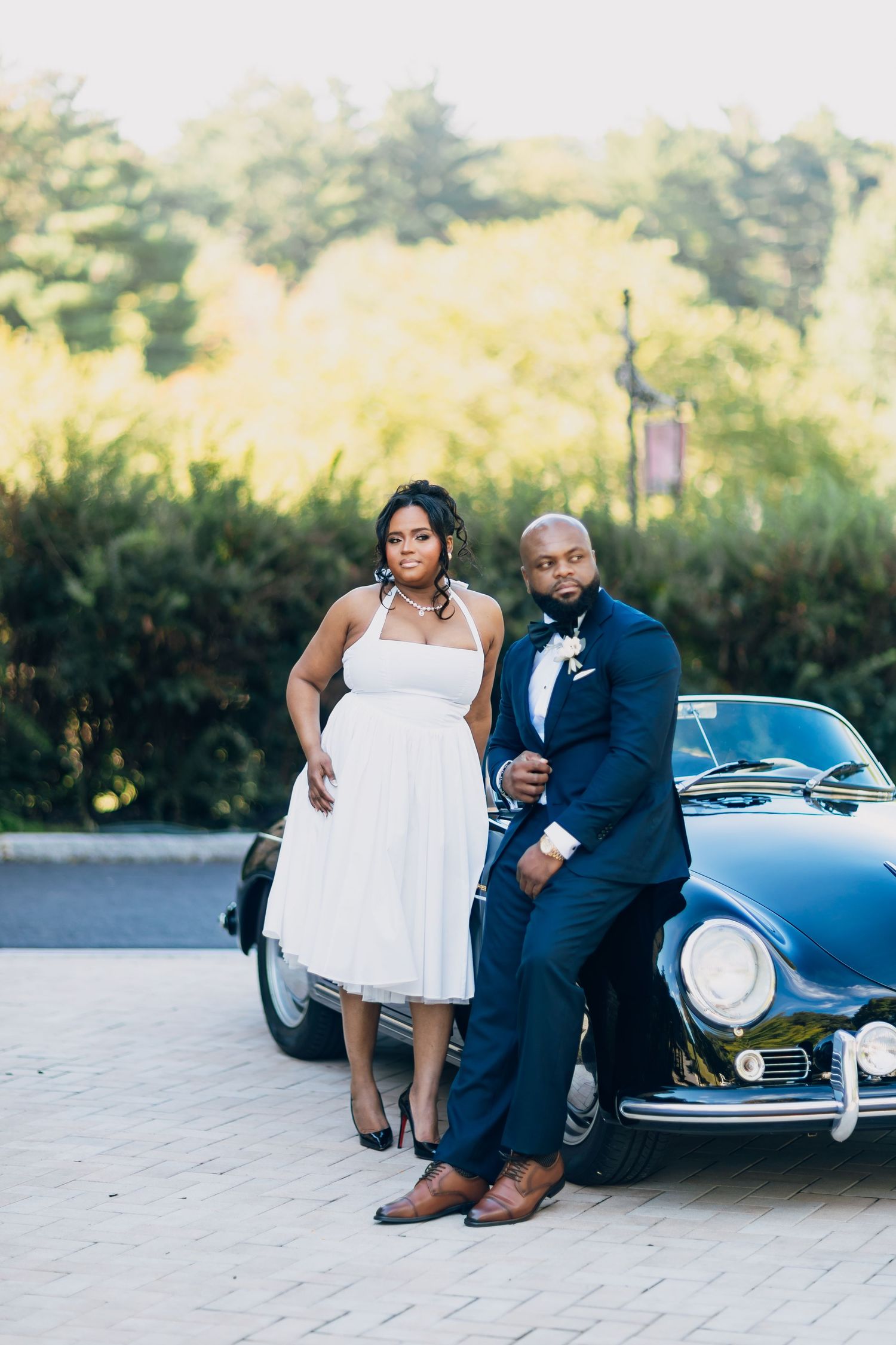 Elegant bride and groom pose together leaning against a classic vintage car on a paved outdoor venue.