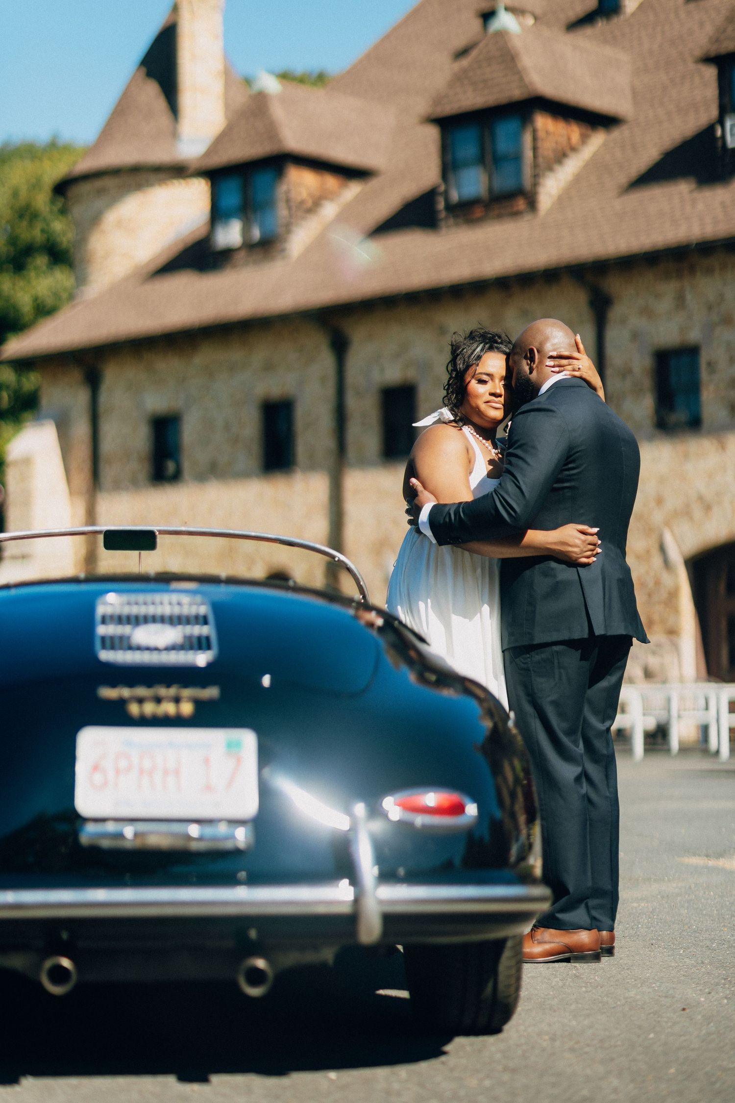 Wedding couple embracing behind a vintage black Porsche in front of a stone castle venue.