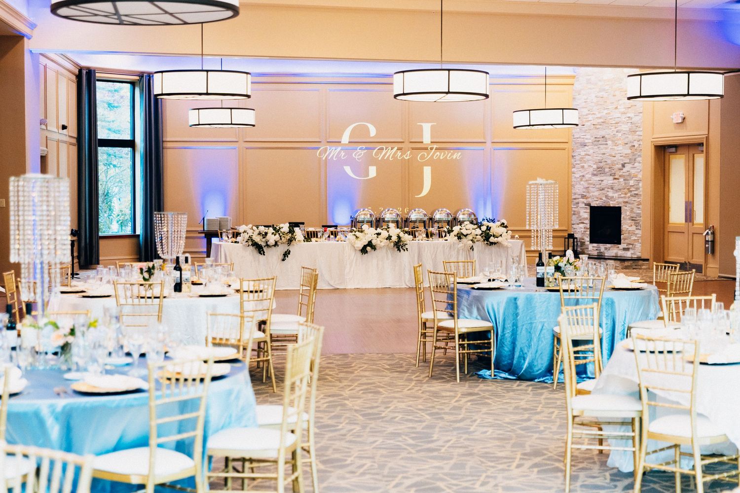 Elegant wedding reception ballroom with blue tablecloths, gold chiavari chairs, floral centerpieces, and a monogram projection on the wall.