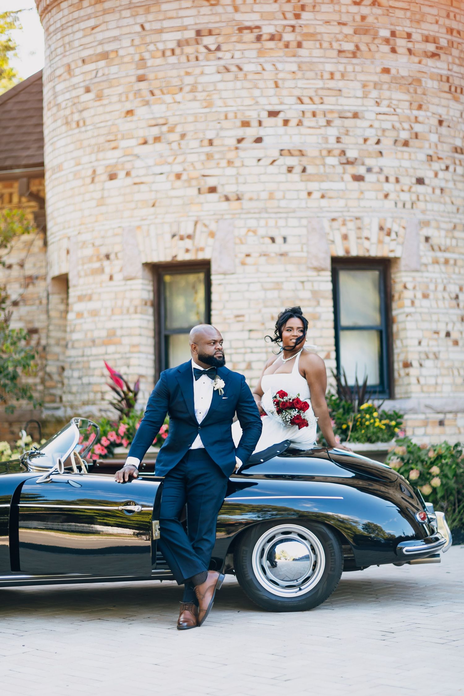 Bride and groom lean against a classic black convertible outside a historic brick tower venue.