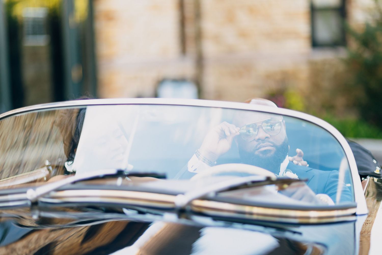 Man sitting in a classic black convertible car, smiling while holding the steering wheel outdoors.