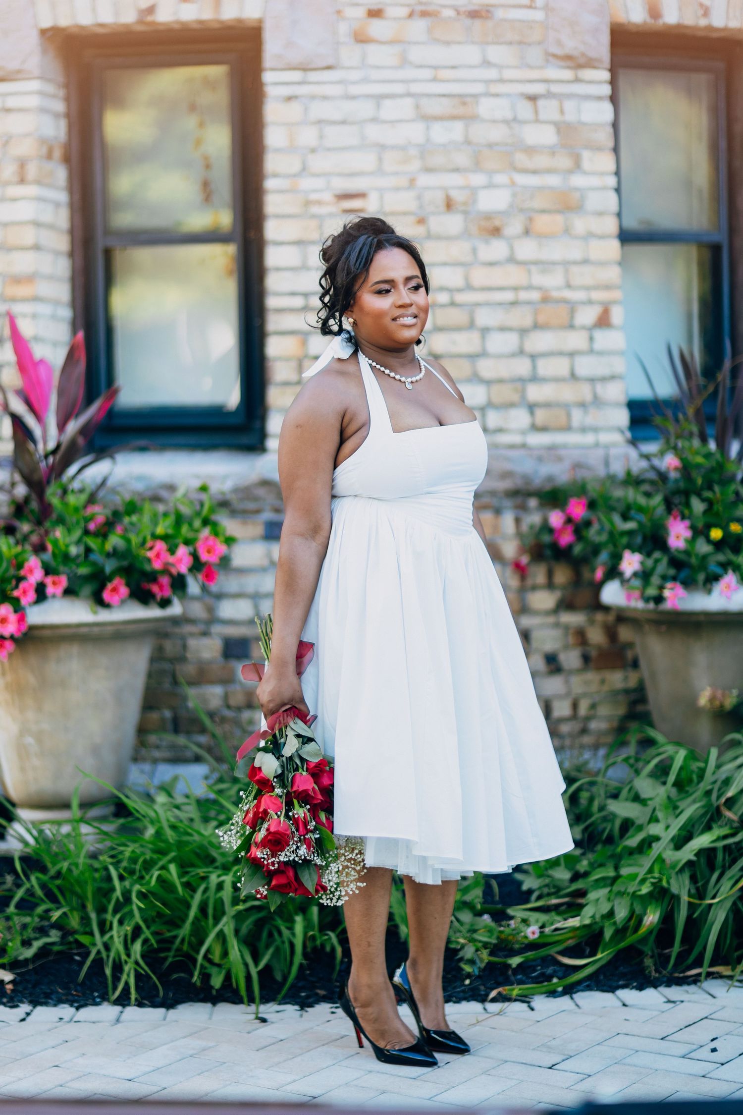 Black bride in white halter midi dress holding red bouquet posing before a brick building.