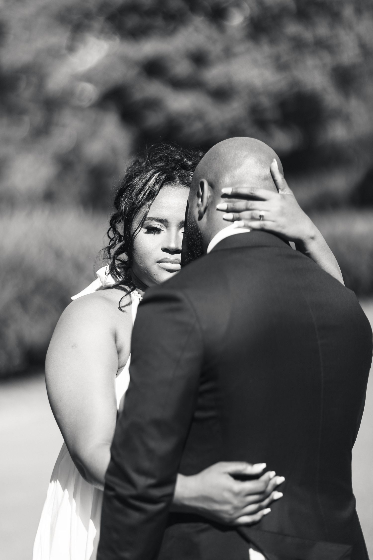 Black and white photo of a bride embracing her groom from behind, gazing softly into the distance.