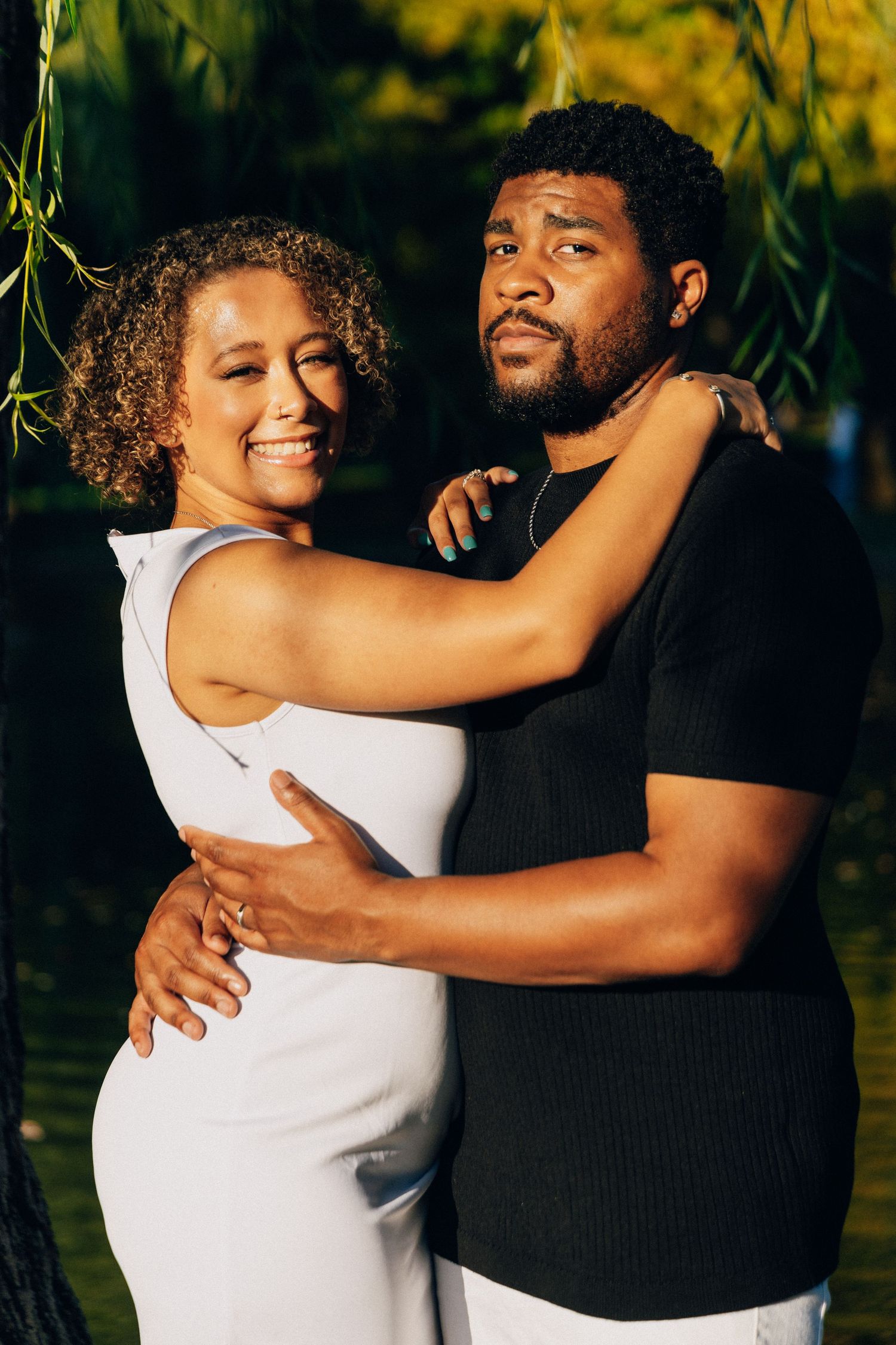 A smiling couple embraces outdoors near a pond, woman in white dress, man in black shirt, surrounded by lush greenery.