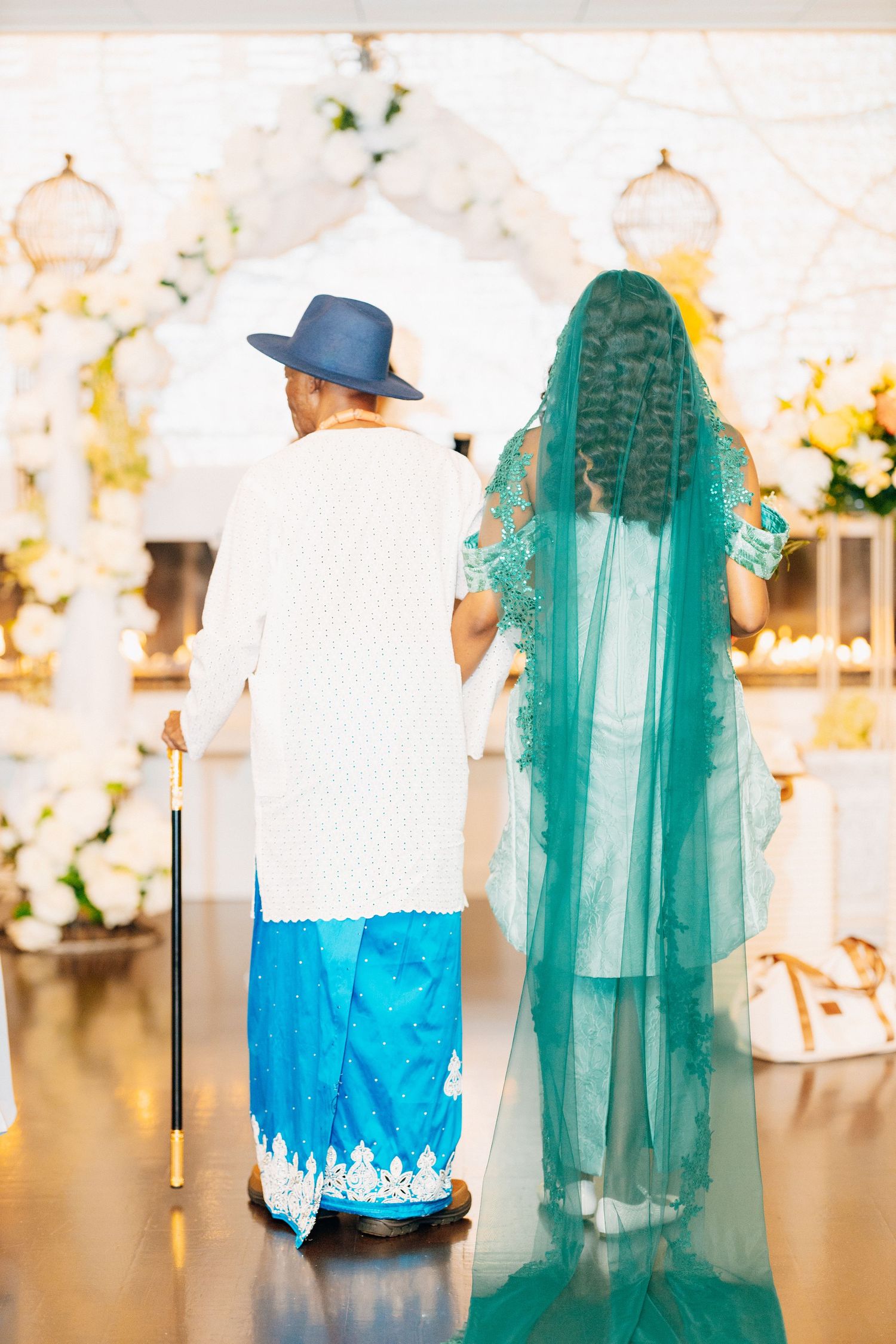 Bride in teal veil walks with elder toward floral wedding arch.