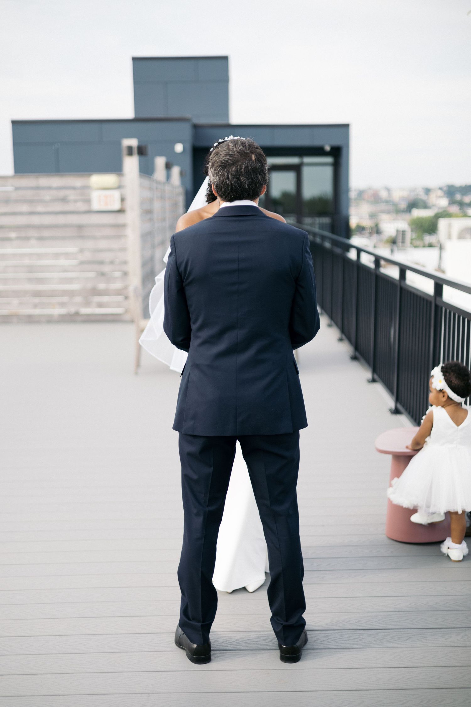 Groom in navy suit shares first look moment with bride on rooftop deck, flower girl nearby.