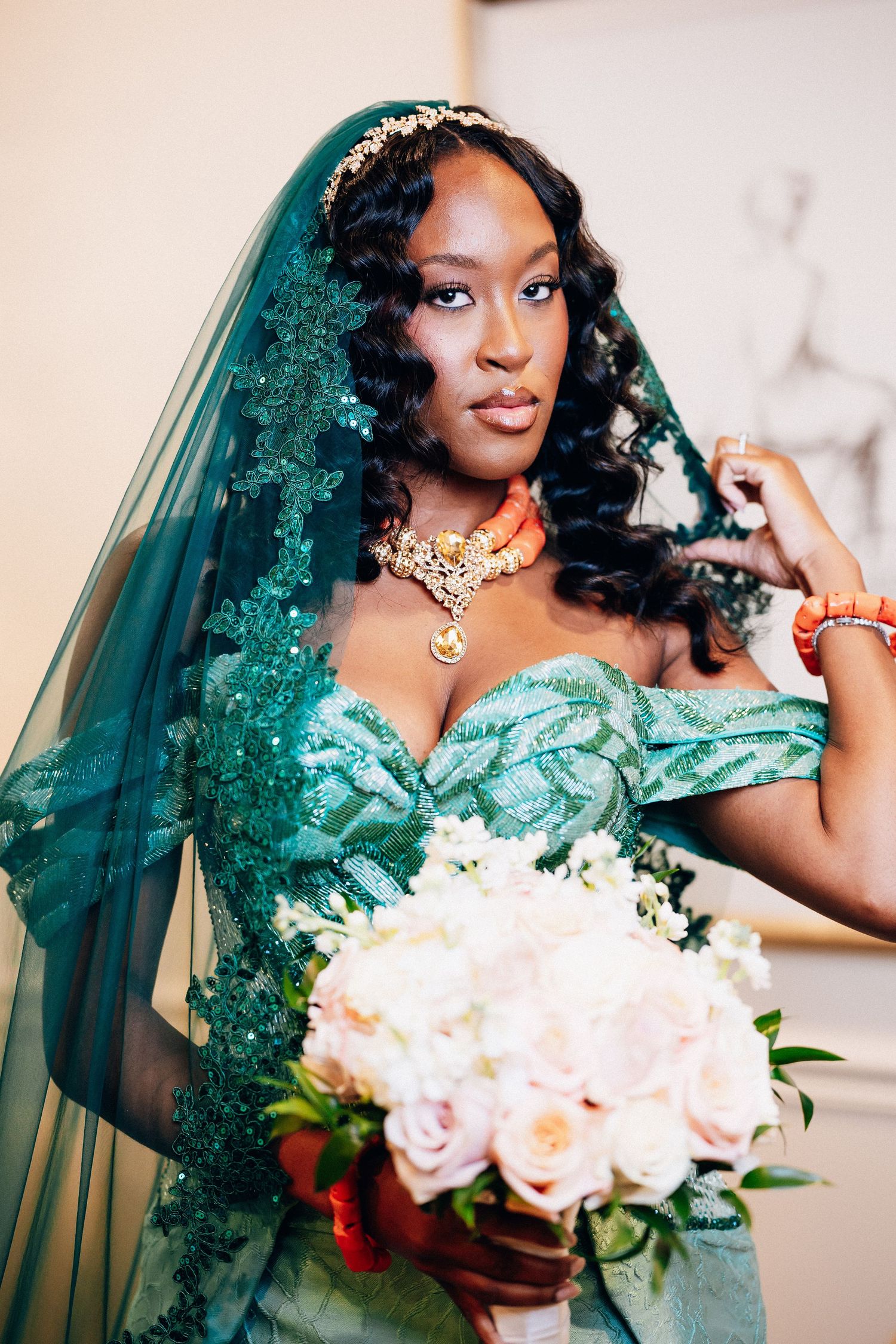 Black bride wearing teal embroidered off-shoulder gown with matching lace veil, coral necklace, holding pink bouquet.