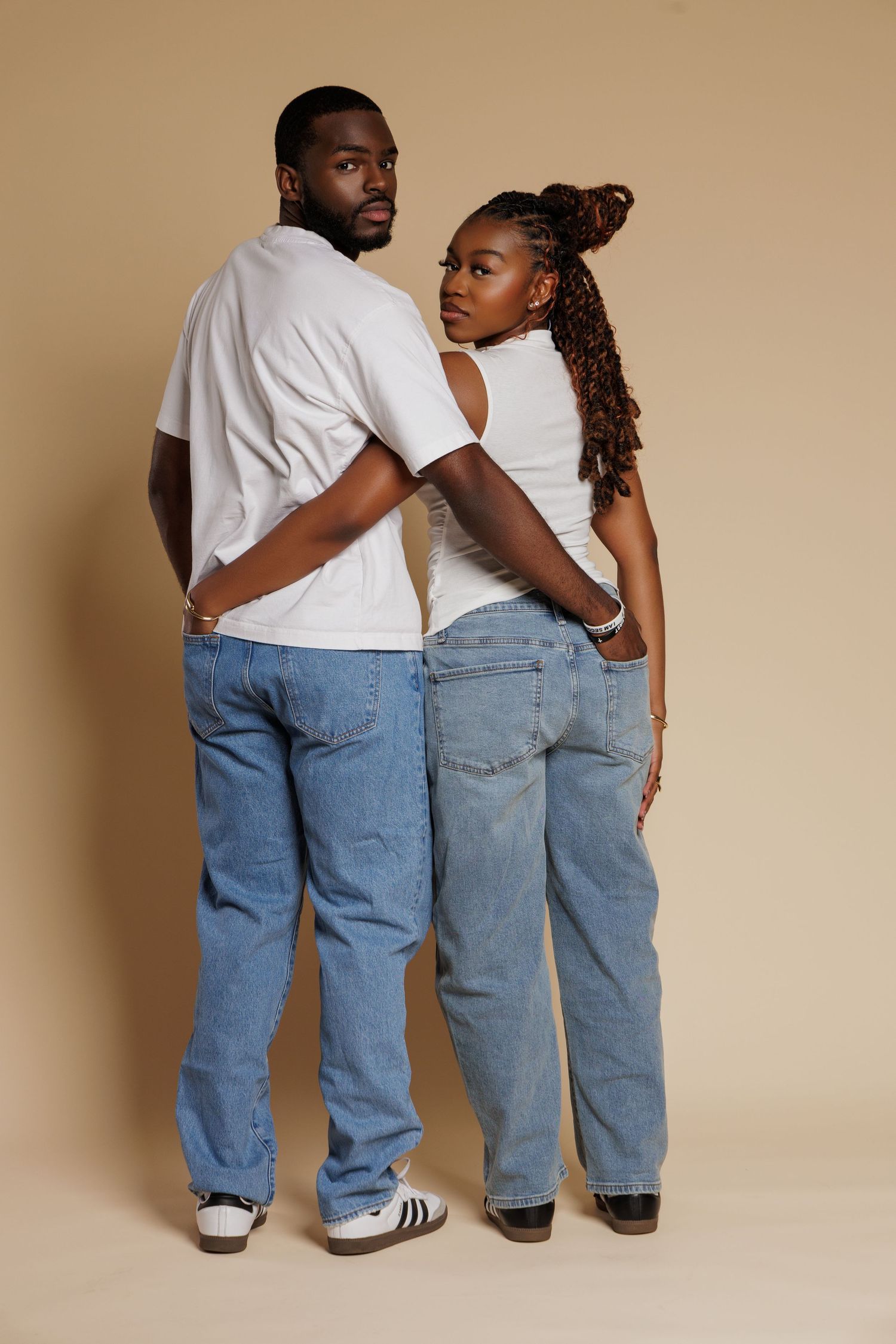 African American couple in matching white tees and blue jeans posing back-to-back on beige background.