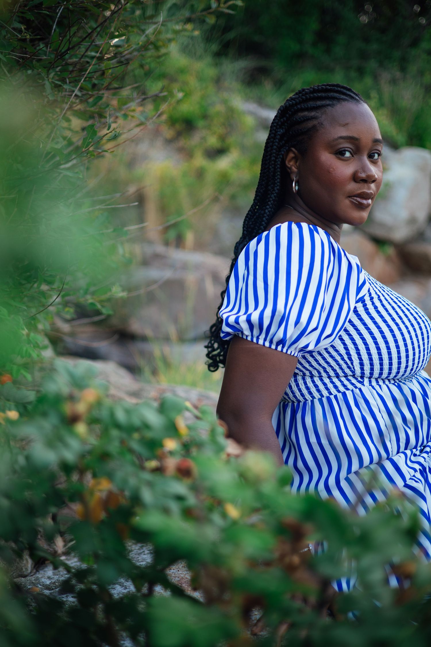 Black woman with braids in blue striped dress posing among green foliage outdoors.