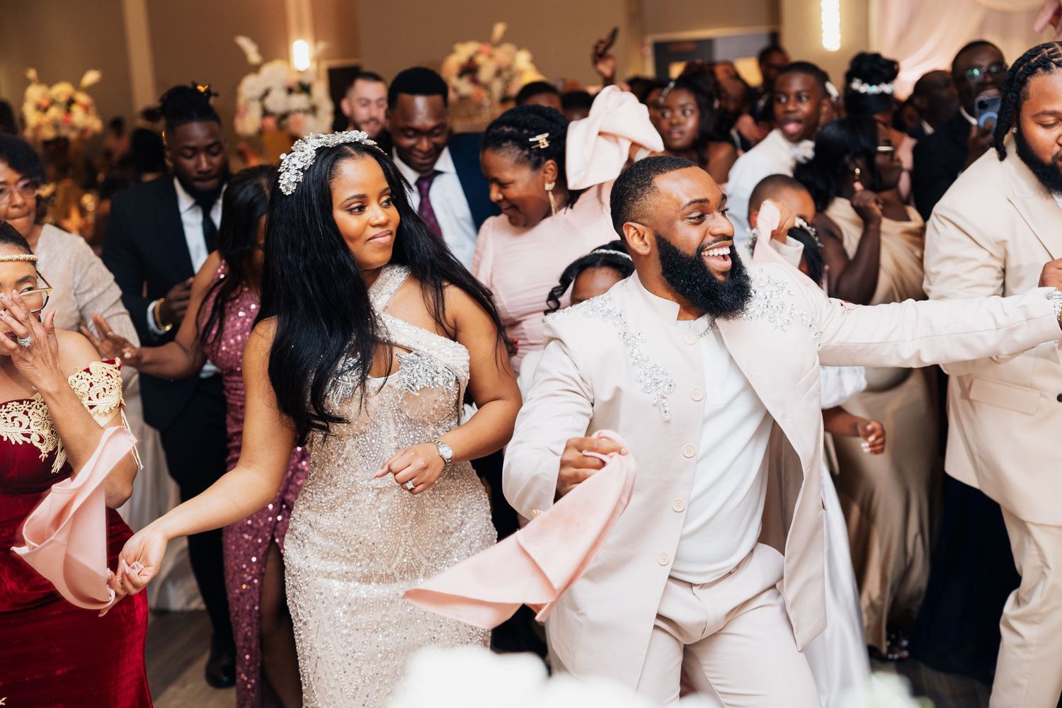 Joyful couple dancing at a wedding reception, groom in white suit, bride in sequin gown, guests celebrating in background.