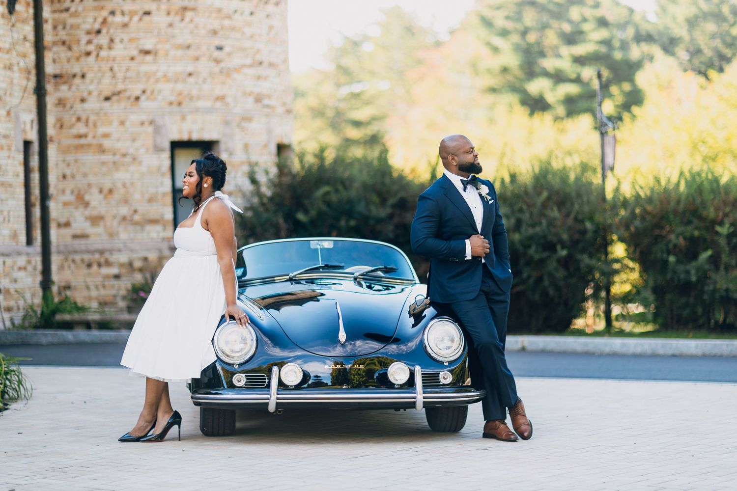 Bride and groom posing elegantly beside a vintage blue Porsche at an outdoor wedding venue.