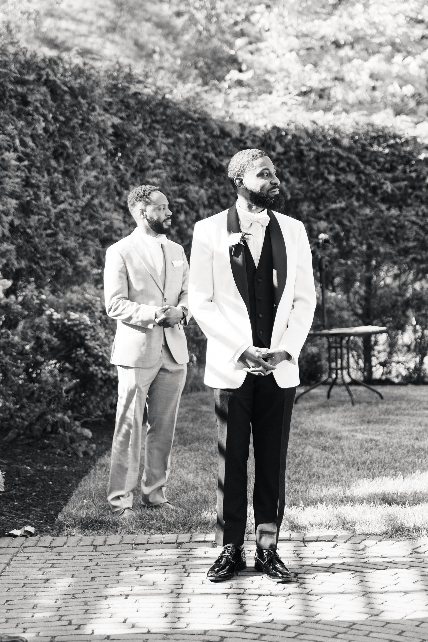 Two grooms in elegant suits stand outdoors in a garden setting on their wedding day.