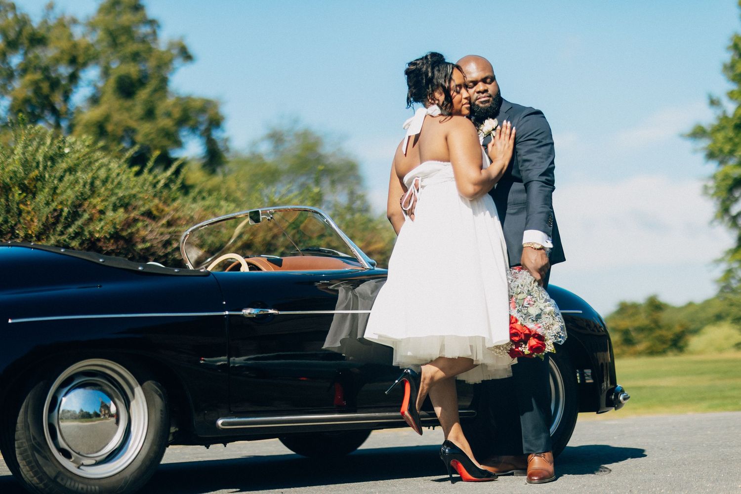 Bride and groom share a romantic embrace beside a classic black convertible on their wedding day.