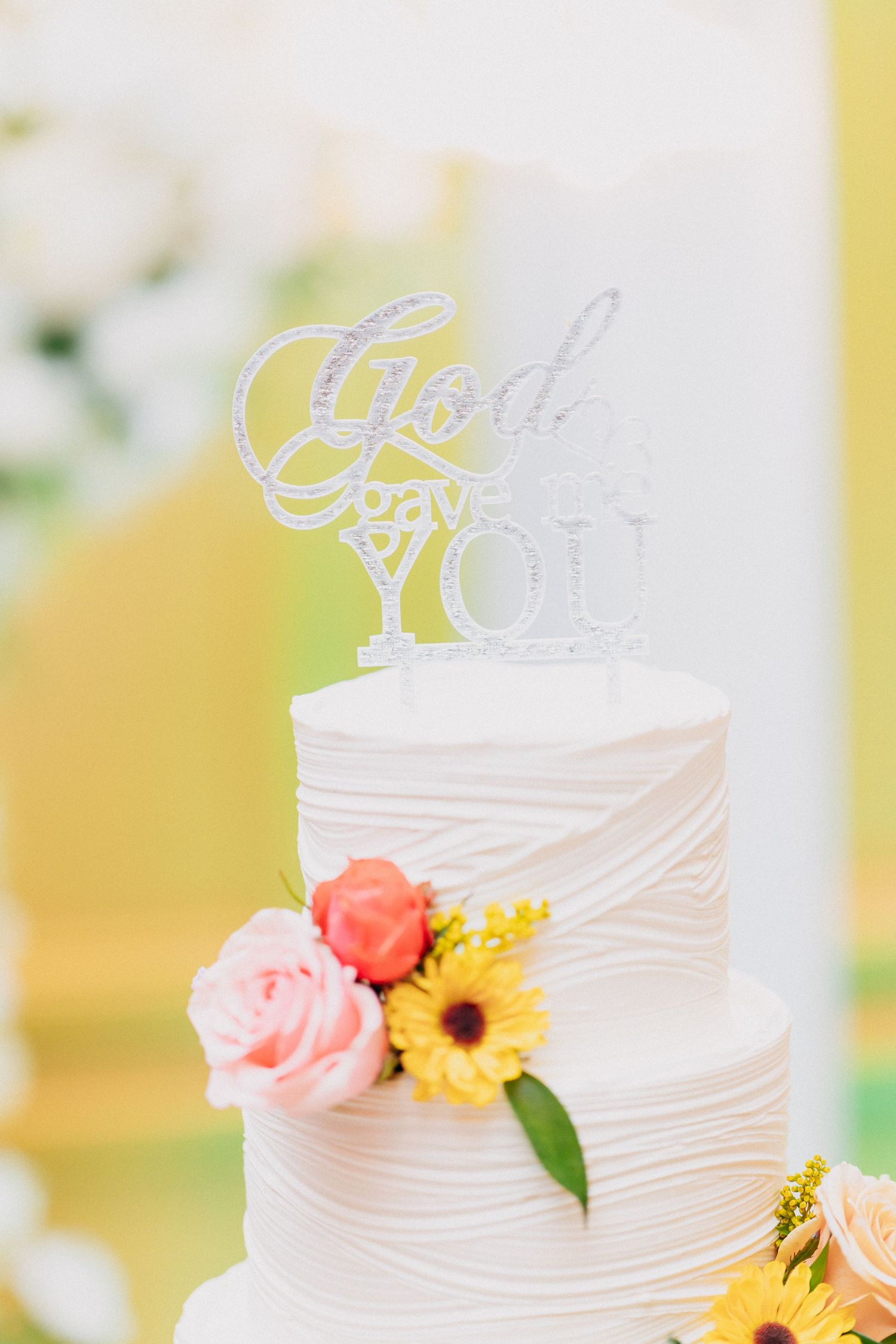 Two-tier white wedding cake with textured frosting, colorful flowers, and a glittery God bless you cake topper.