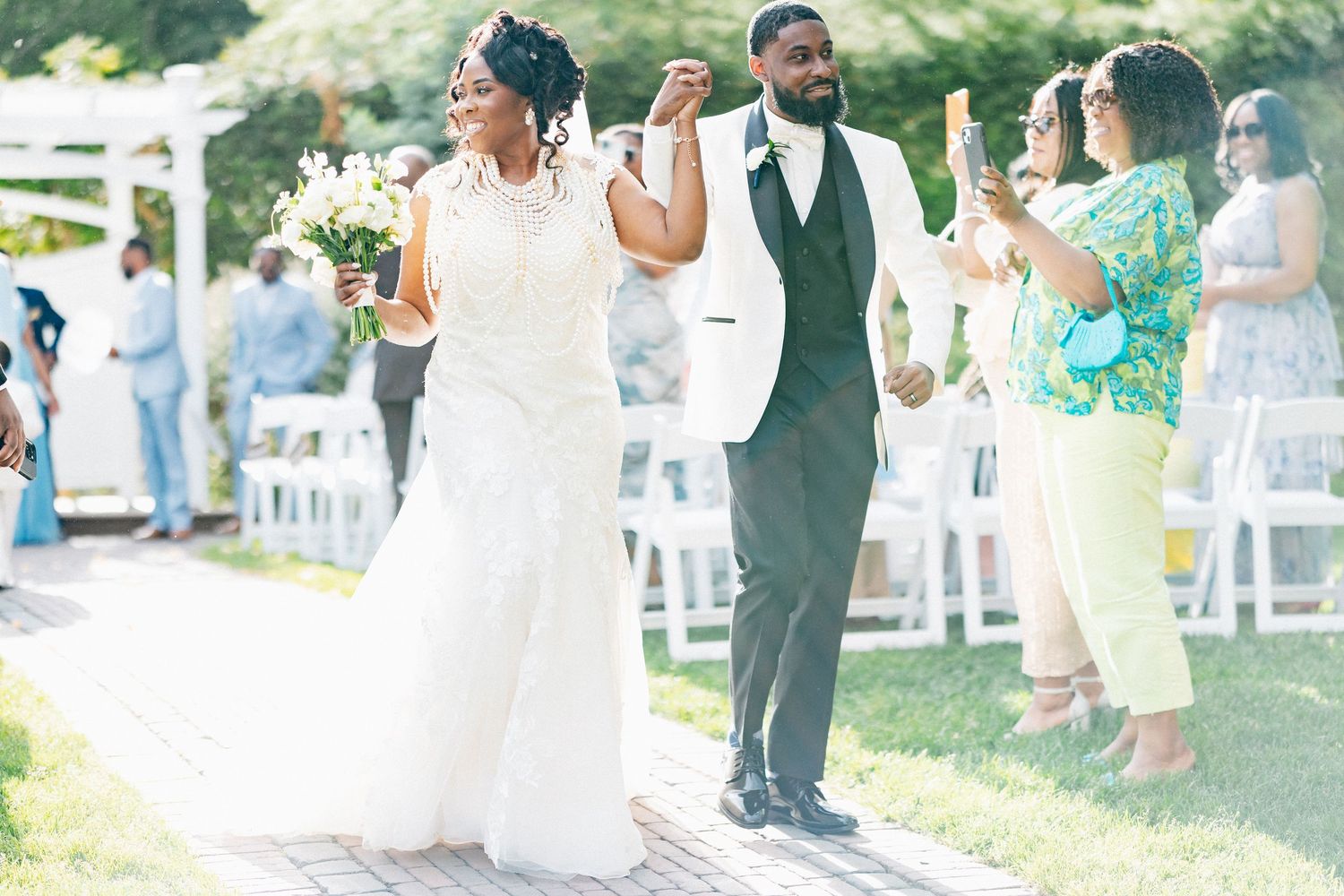 Joyful newlywed couple walks down the aisle after outdoor wedding ceremony, bride in white gown, groom in white jacket.