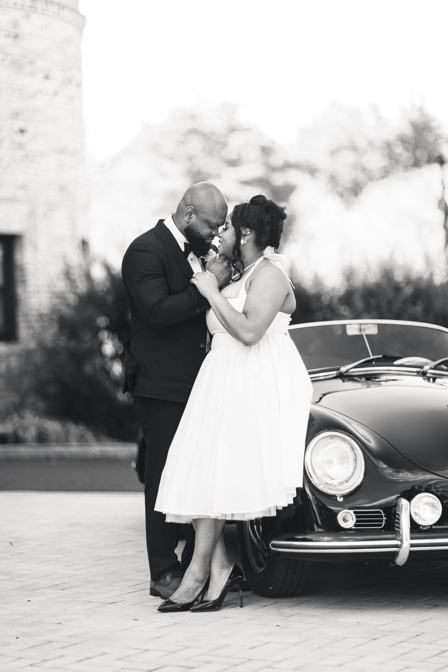 Black and white photo of bride and groom sharing romantic moment beside vintage Porsche.