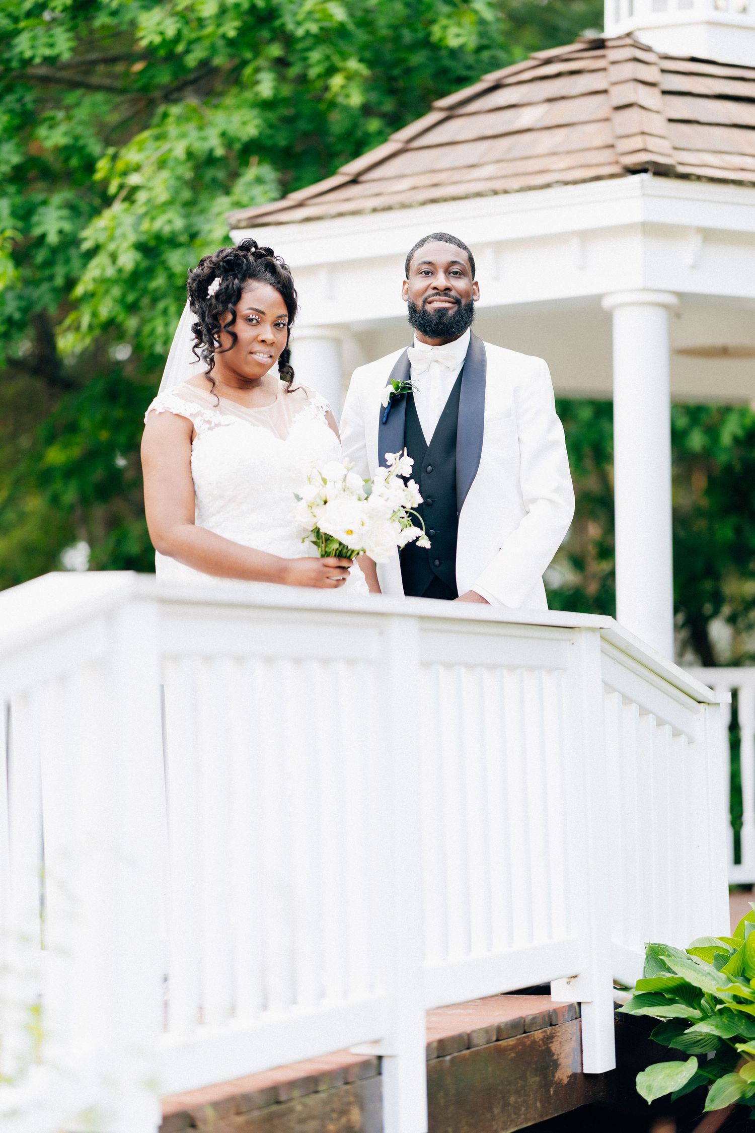 A Black bride and groom pose at a white gazebo, bride holding white flowers in lace dress, groom in white tux.