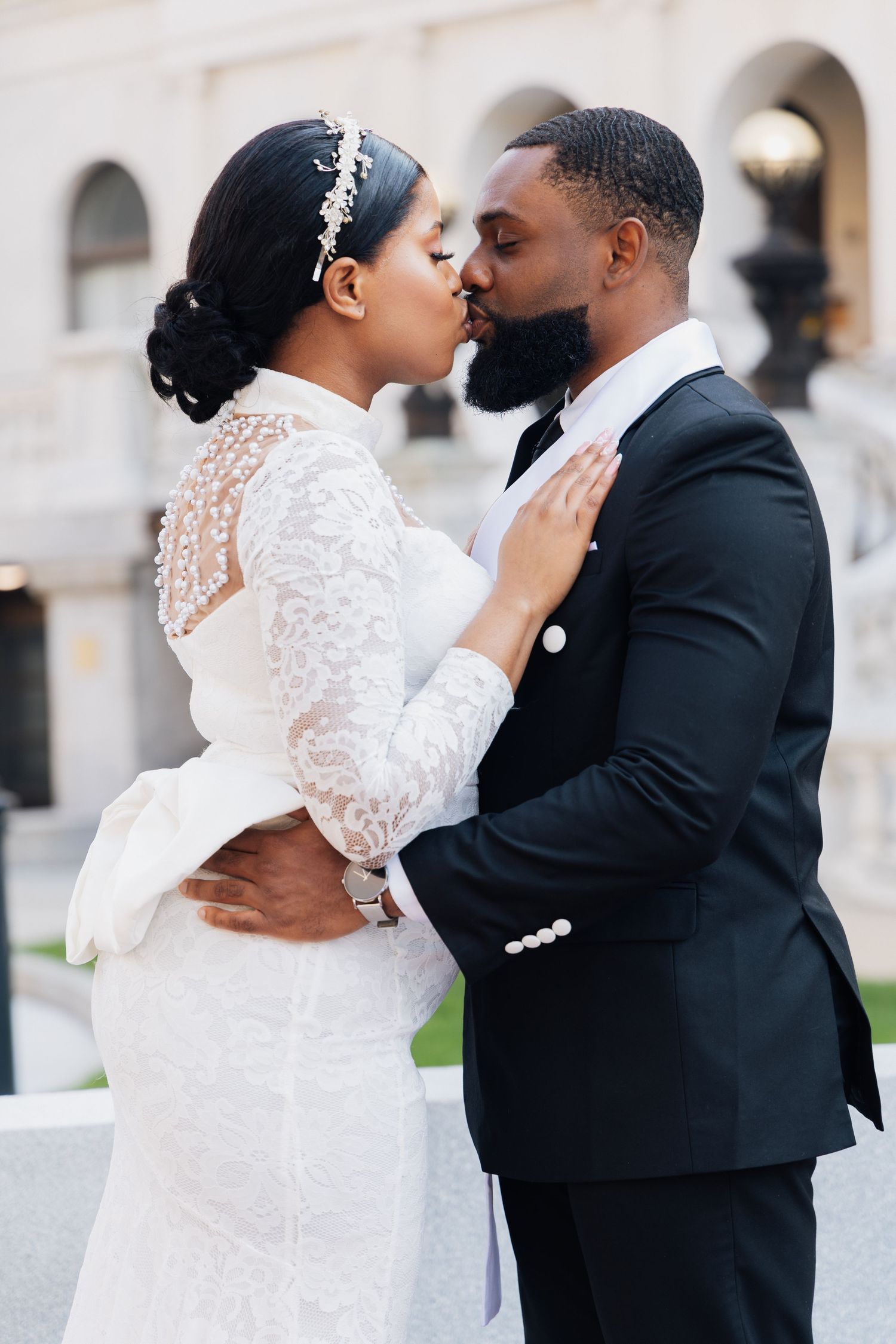 A Black bride and groom share a romantic kiss outdoors, she in white lace gown, he in classic black tuxedo.
