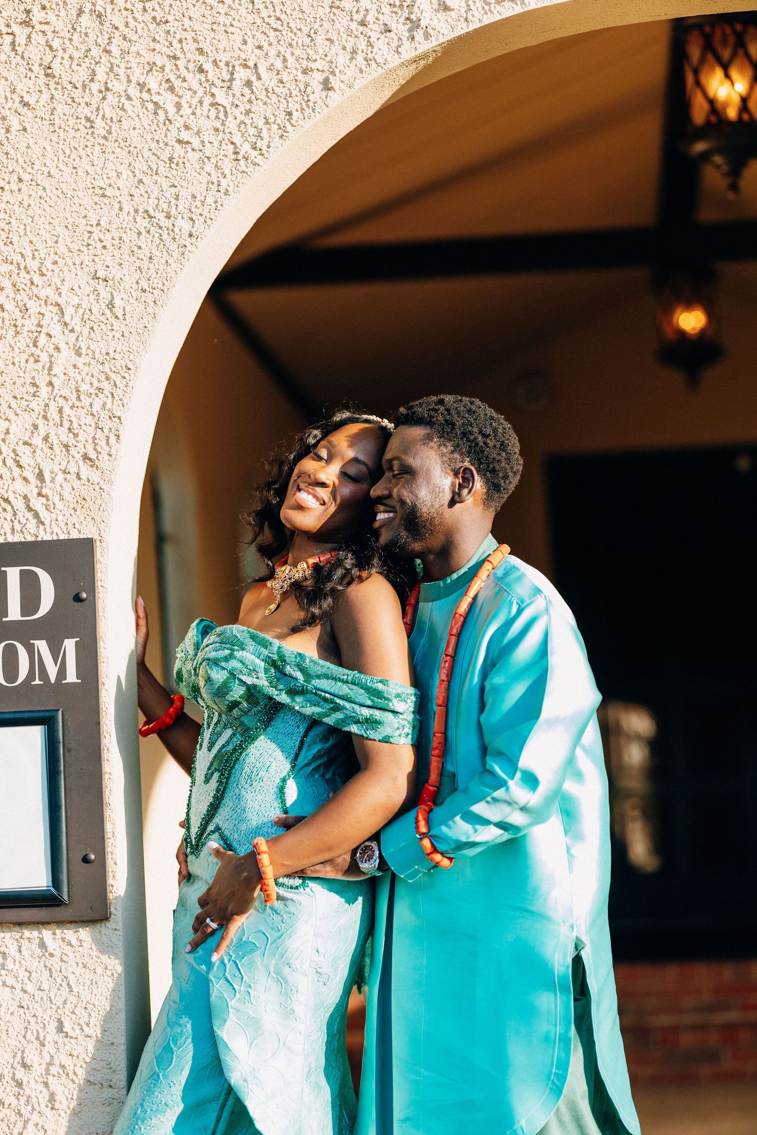 African couple in matching teal traditional attire embrace under an archway, smiling joyfully.