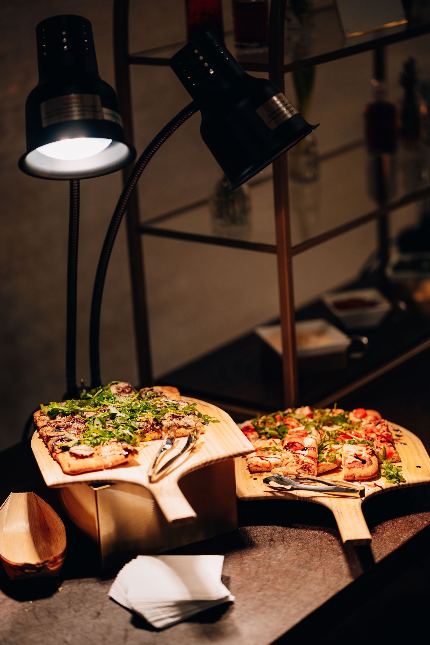Two pizzas on wooden boards illuminated by black heat lamps at food station.