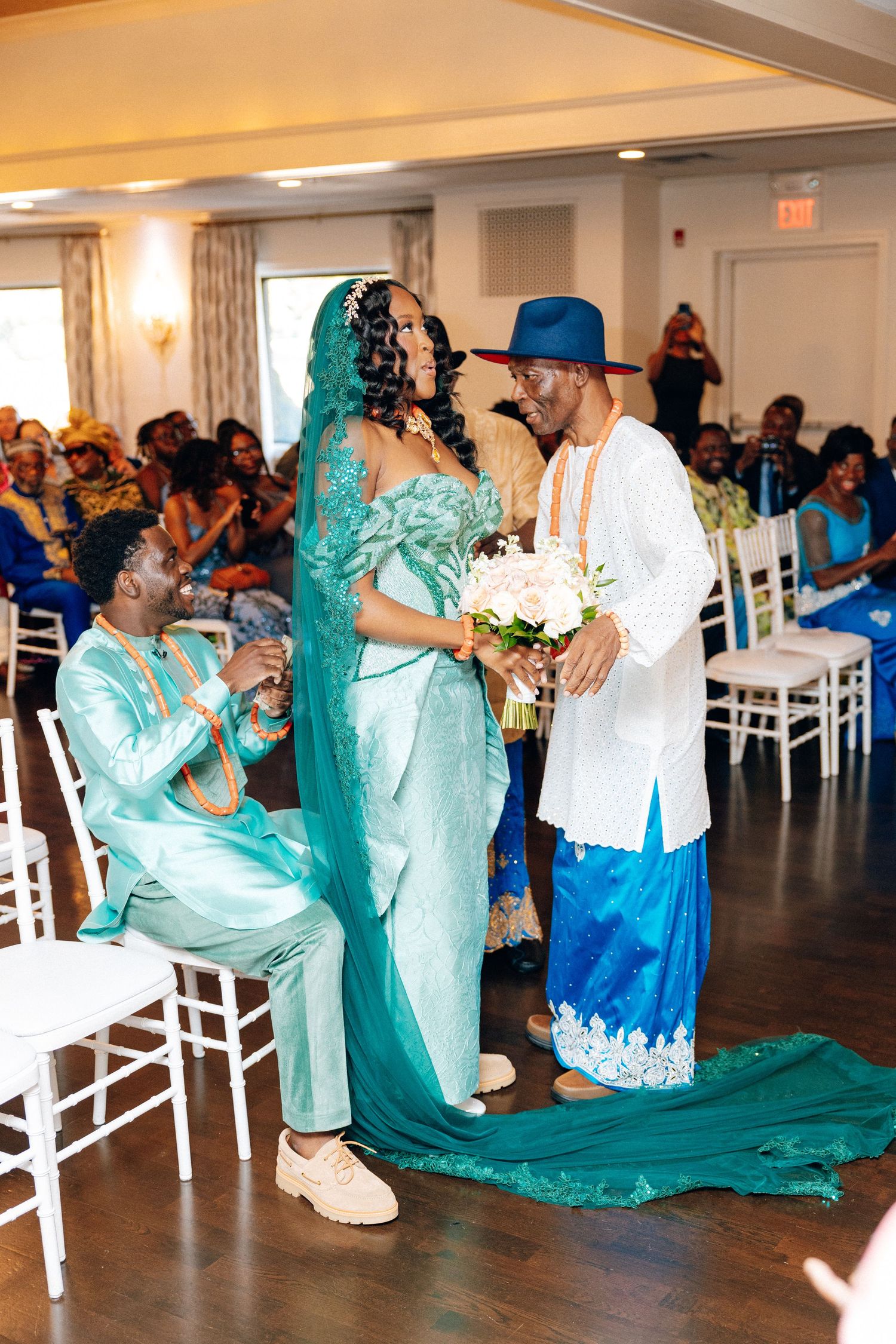 Bride in teal gown stands with two men in African attire during traditional wedding ceremony.