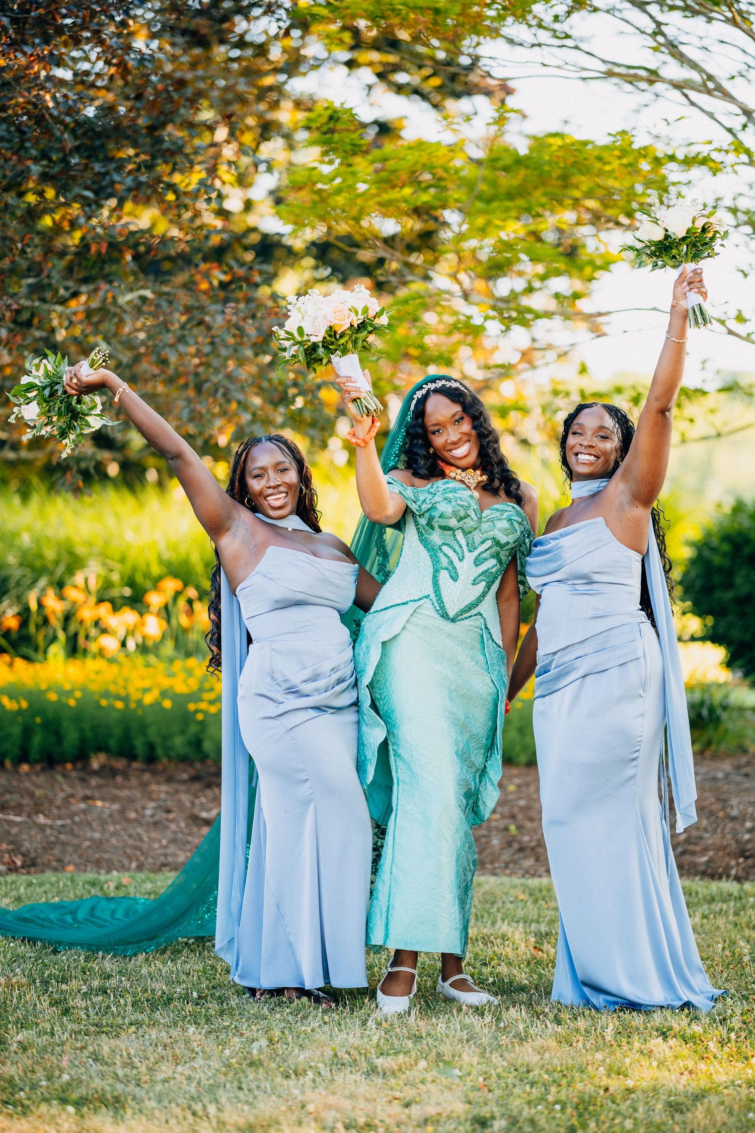 Bride and two bridesmaids in blue dresses celebrate joyfully raising bouquets in sunny garden setting.