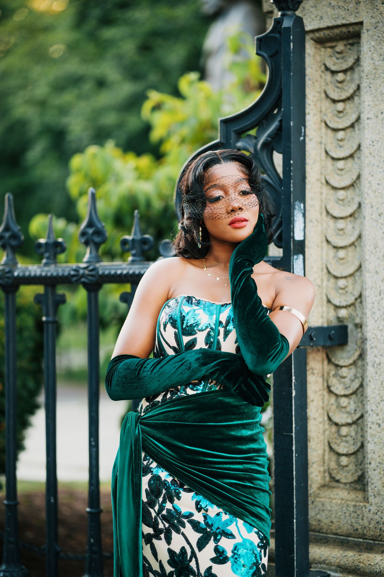 A woman in a green floral strapless gown with velvet gloves and veil poses elegantly by ornate iron gates.