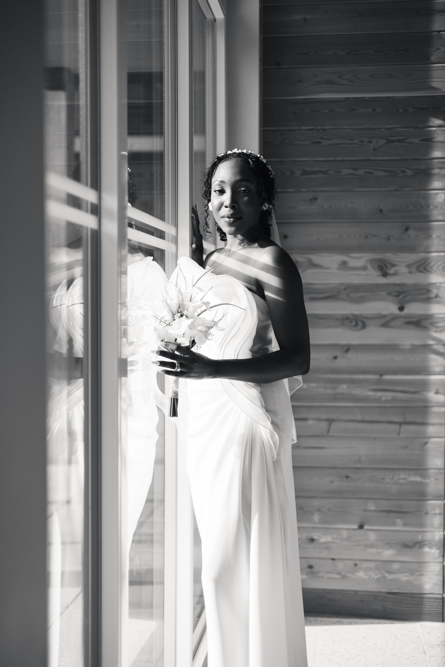 Black and white portrait of African American bride in white gown holding bouquet beside window with veil.