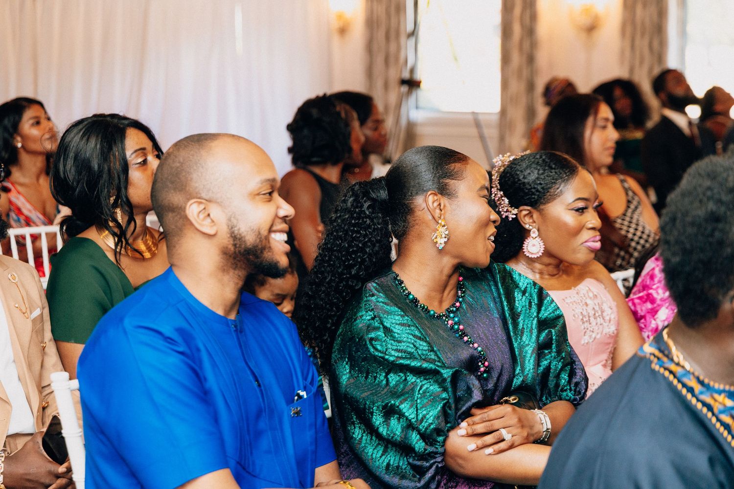Guests in colorful traditional attire smile and laugh together at a wedding ceremony celebration.