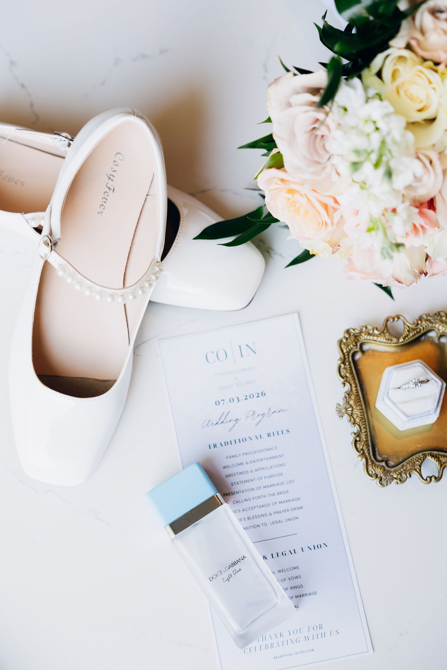 White bridal heels with pearl straps beside a floral bouquet, wedding program, perfume, and gold ring dish on marble.