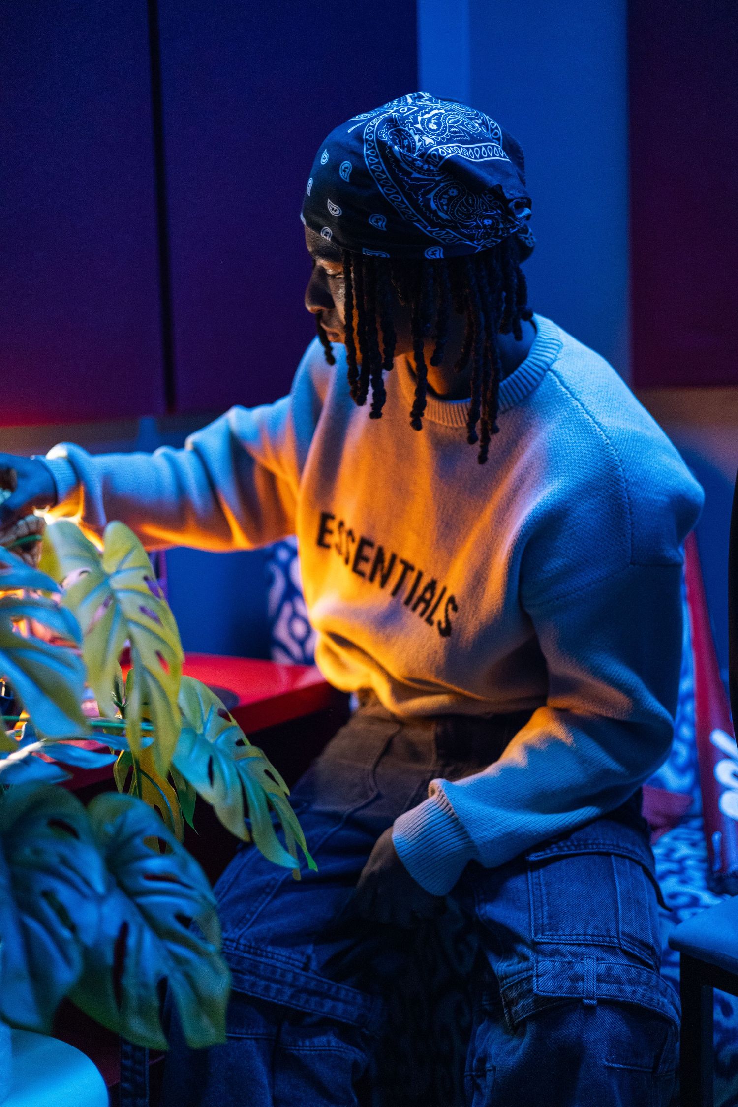 Young man with dreadlocks wearing Fear of God Essentials sweatshirt and bandana in neon-lit studio.