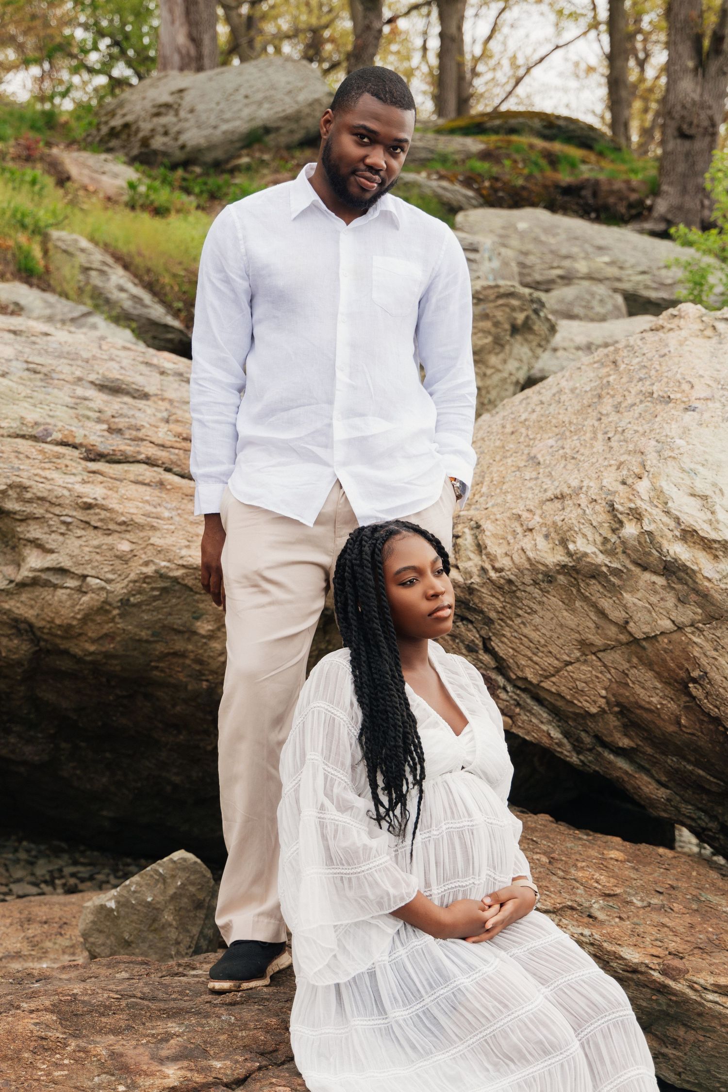 Pregnant Black woman in white dress sits on rocks with partner in white shirt standing behind her outdoors.