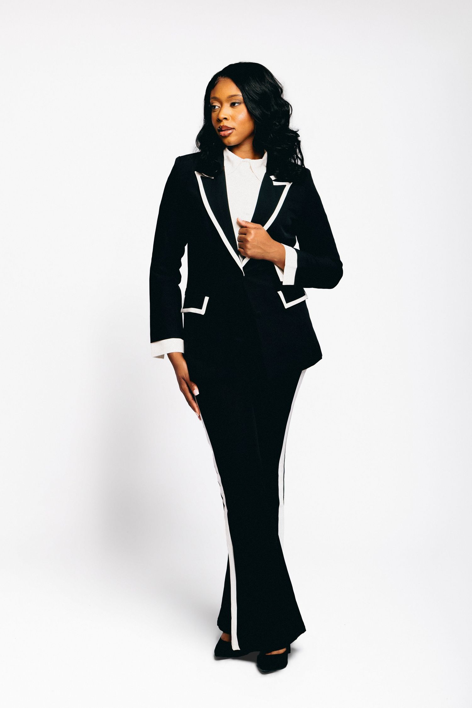 Woman in black and white tuxedo suit with contrast trim, posing confidently against white background.