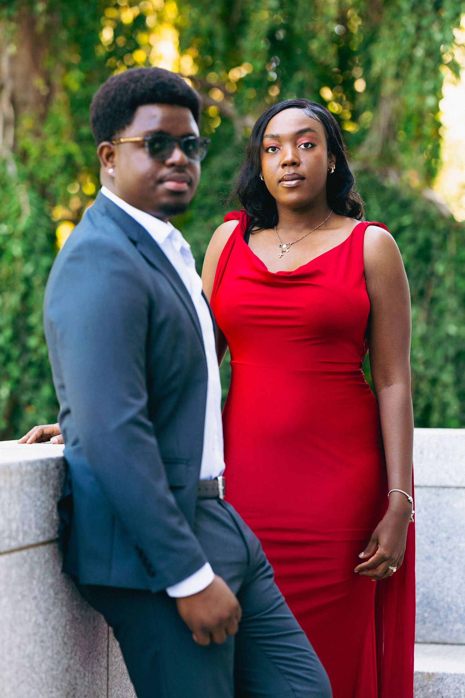 A young couple dressed in formal attire, man in gray suit and woman in red gown, posing outdoors near stone wall.