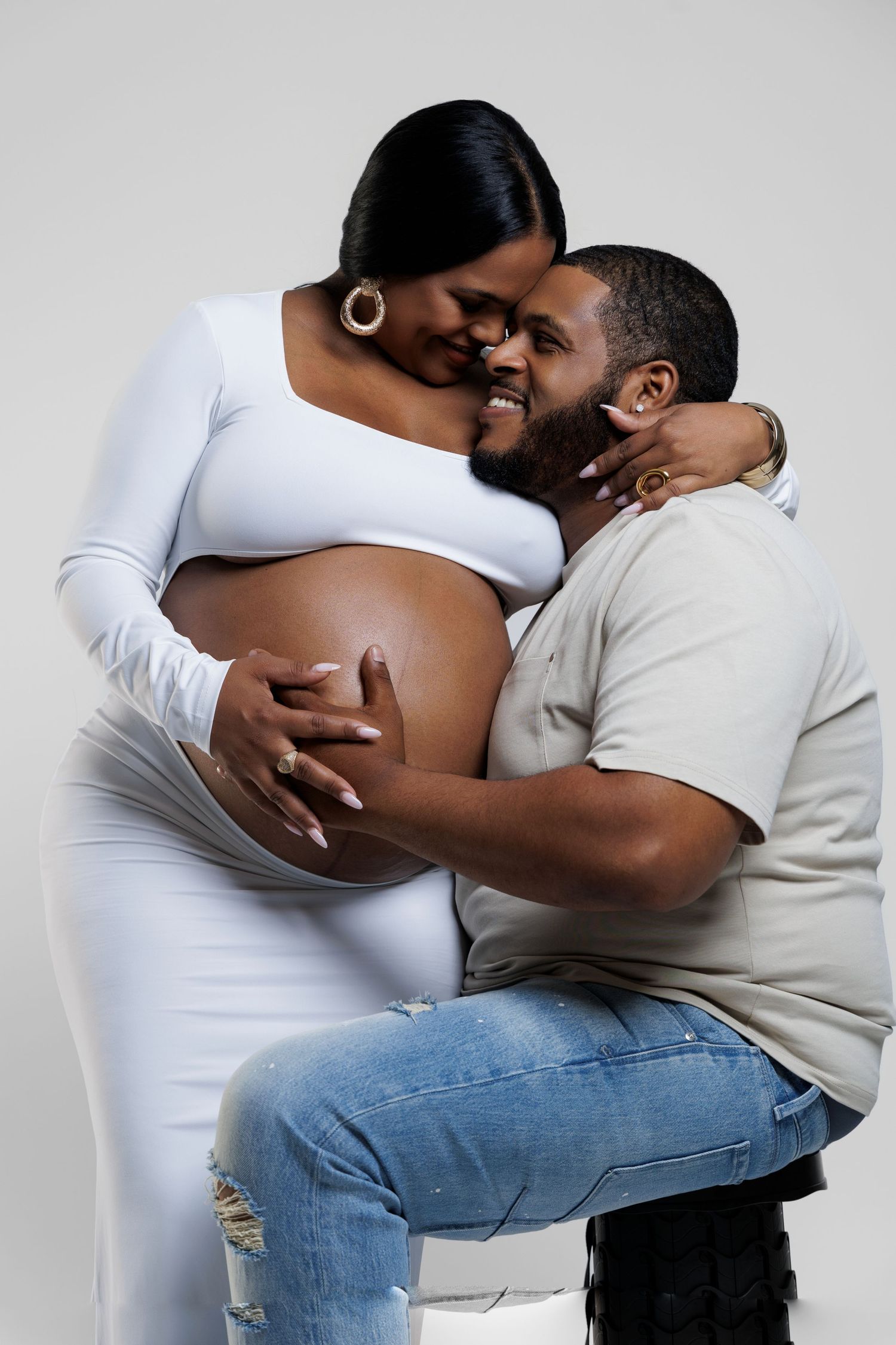 A pregnant Black woman in a white dress holds her baby bump while her partner kisses her cheek during a maternity photoshoot.