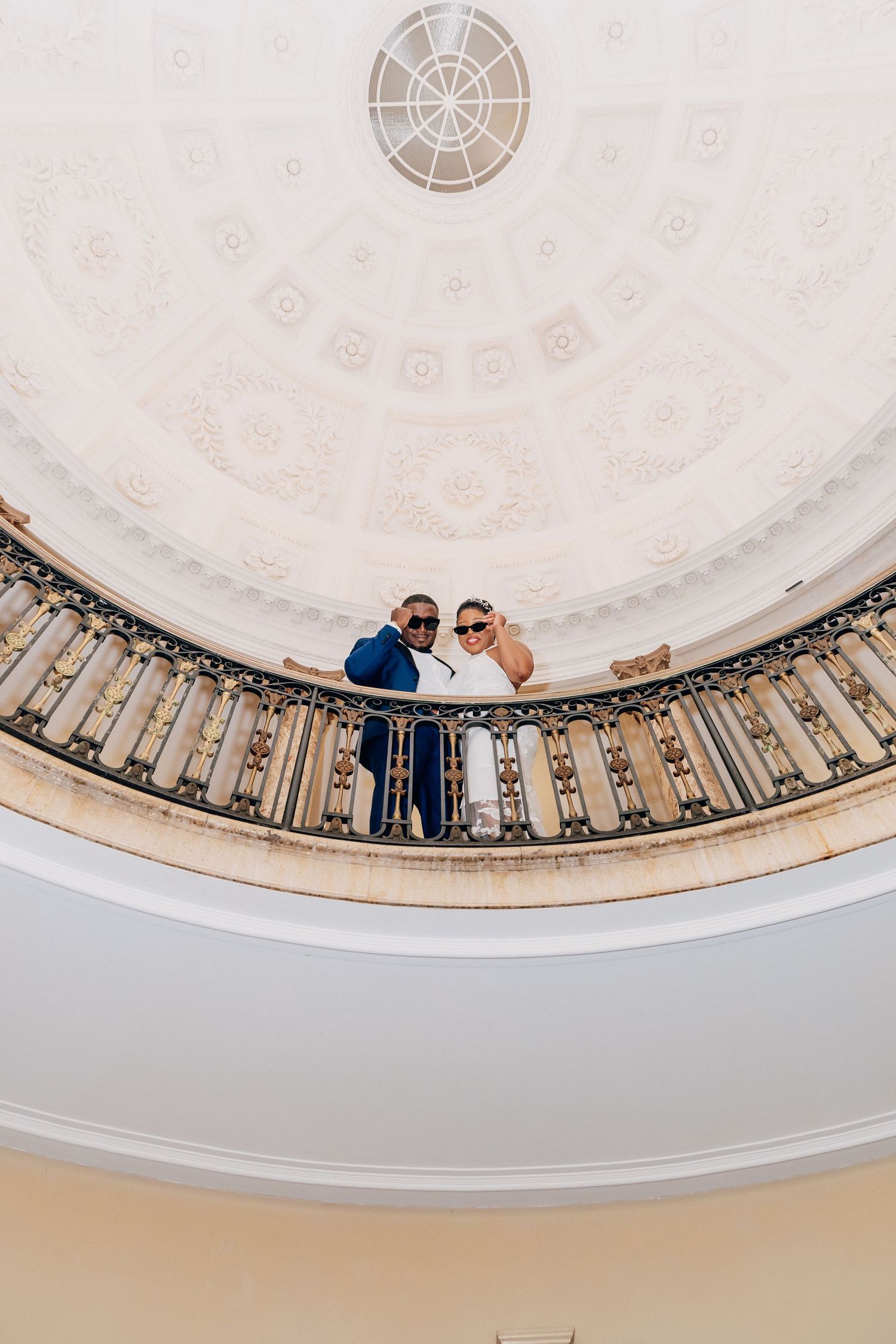 Couple in wedding attire posing on ornate curved balcony beneath elegant domed ceiling.