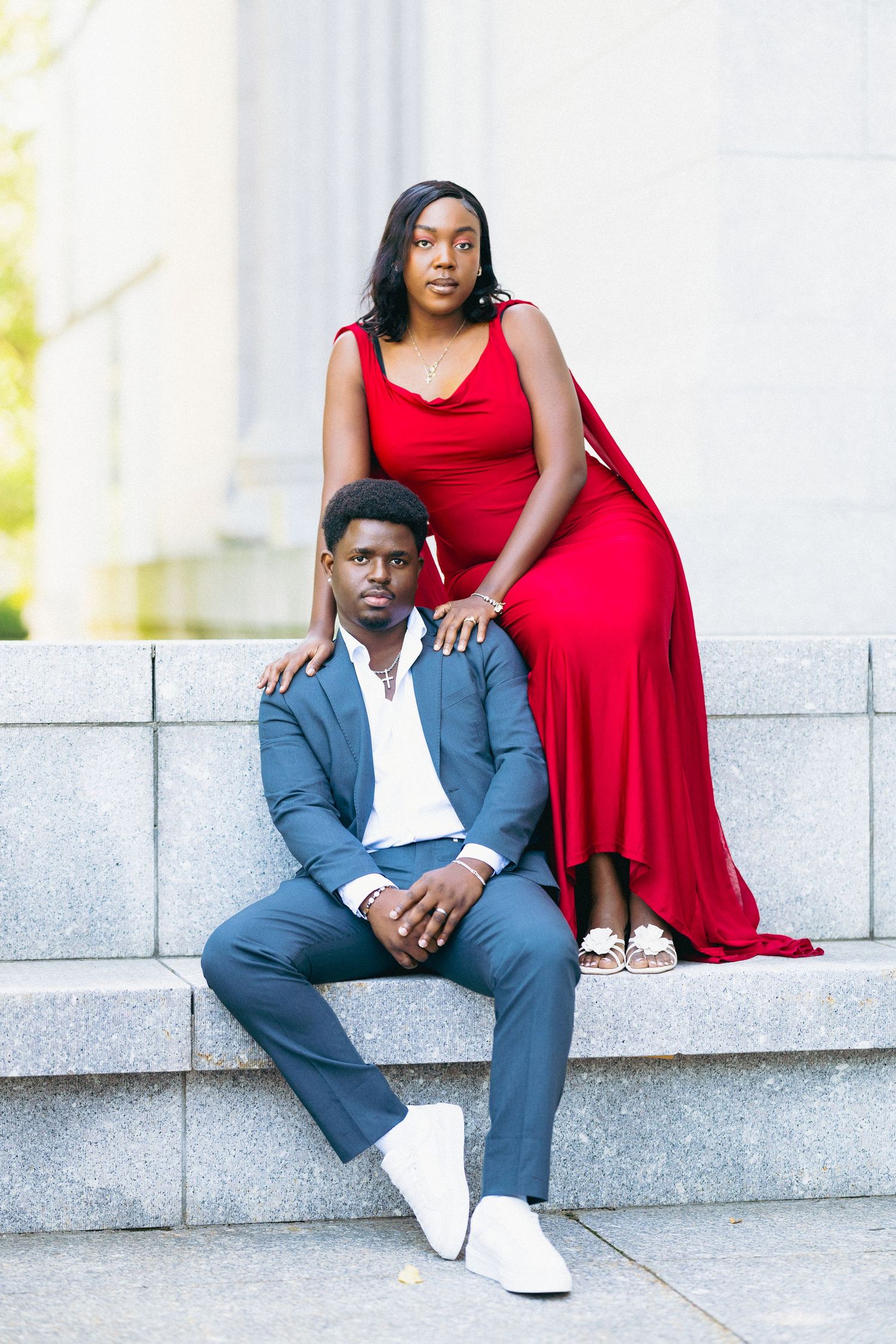 Elegant Black couple posing on stone steps, woman in red gown, man in blue suit with white sneakers.