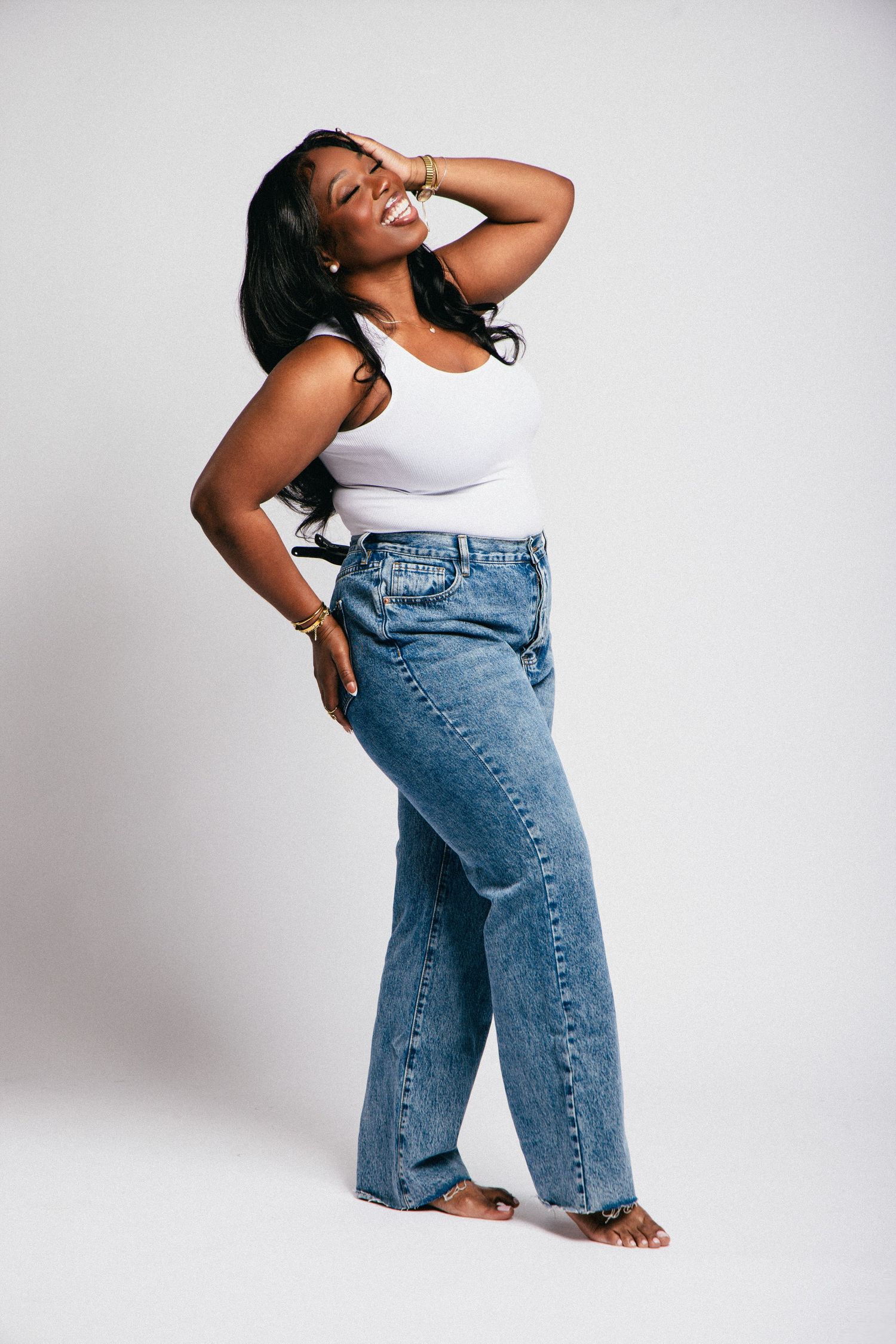 Smiling Black woman in white tank top and wide-leg blue jeans posing confidently against white background.