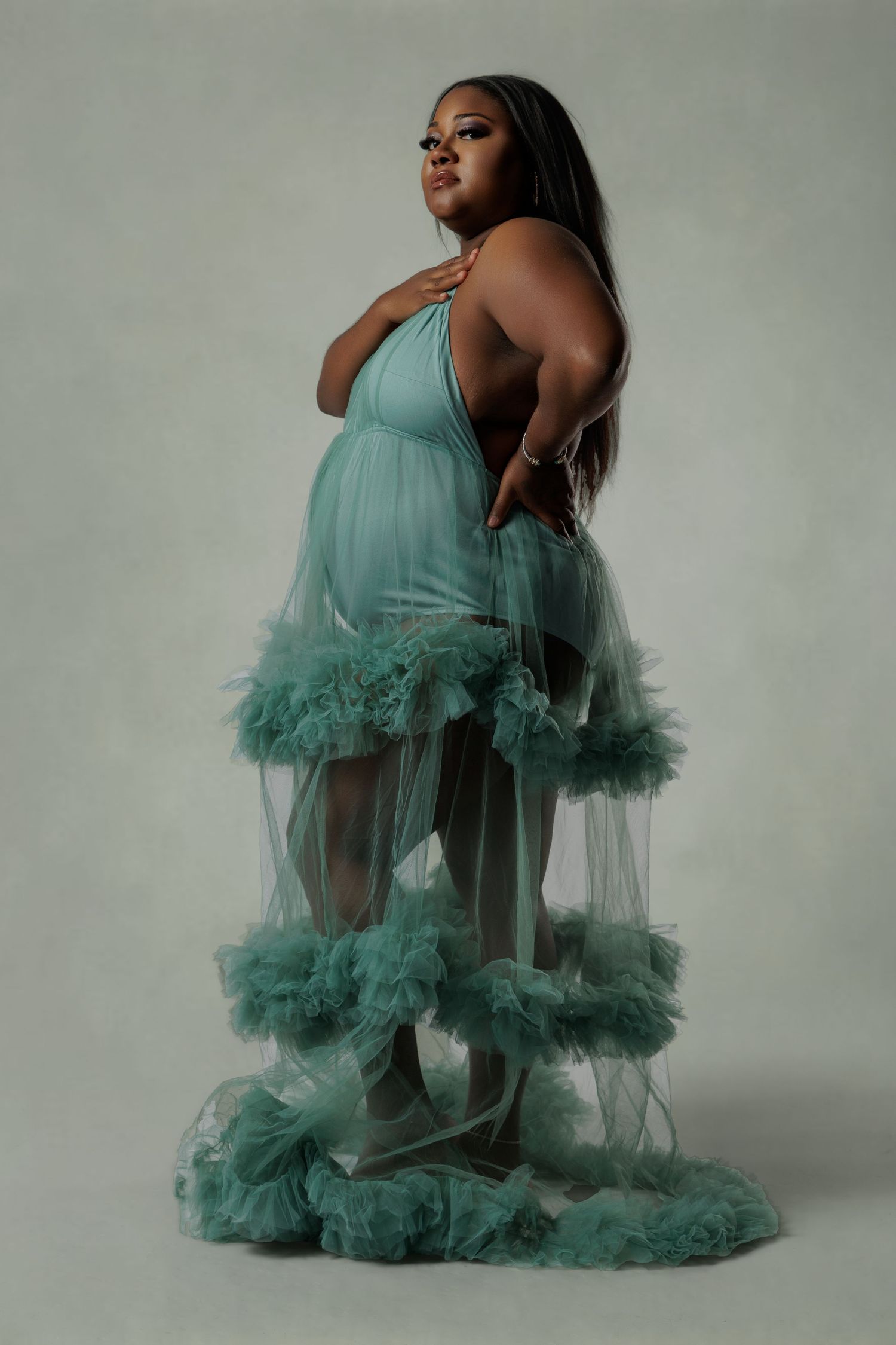 Pregnant woman in elegant teal ruffle tulle maternity gown posing gracefully in a studio photo shoot.