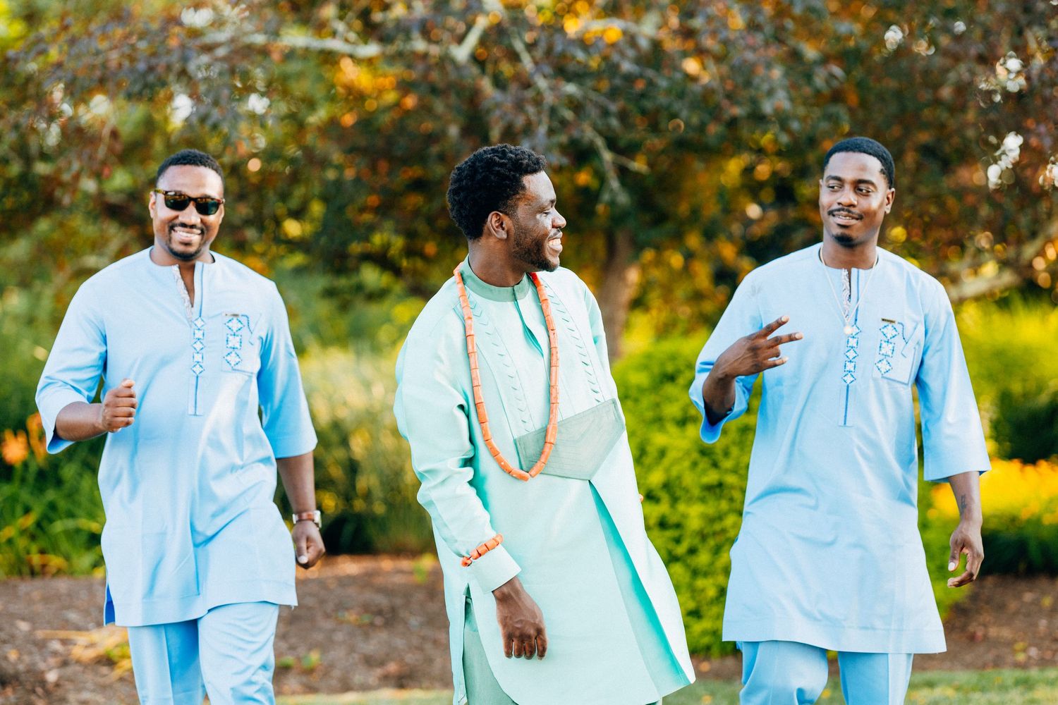 Three men in traditional African attire walk outdoors, wearing light blue and mint green outfits with orange beaded accessories.