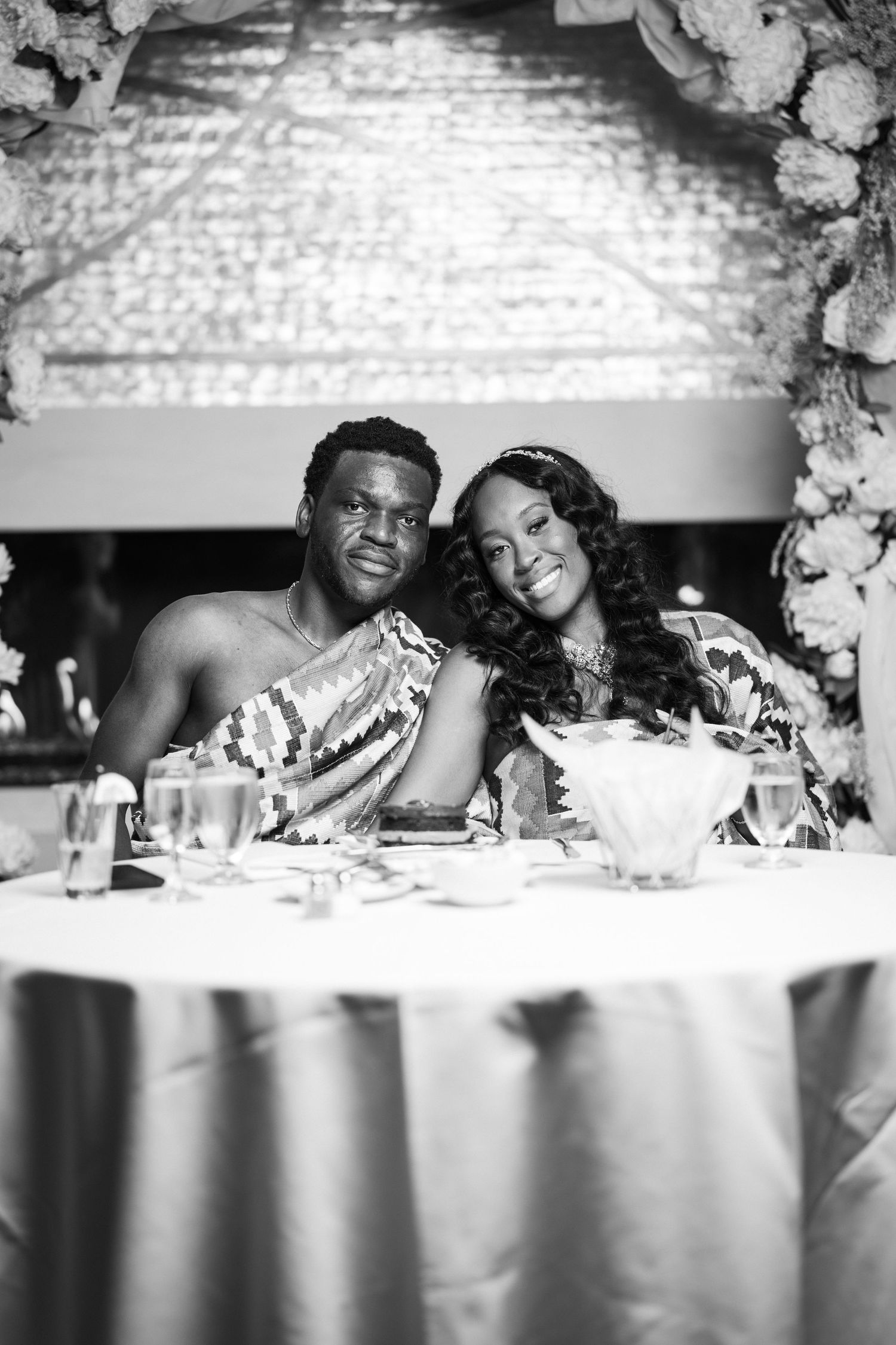 Black and white portrait of a couple in traditional African attire seated at a decorated sweetheart table.