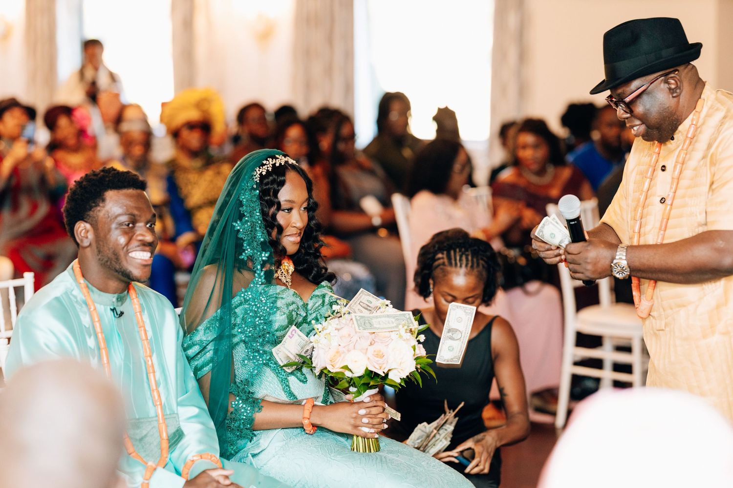Bride and groom in matching teal traditional African attire receive blessings with money at Nigerian wedding ceremony.