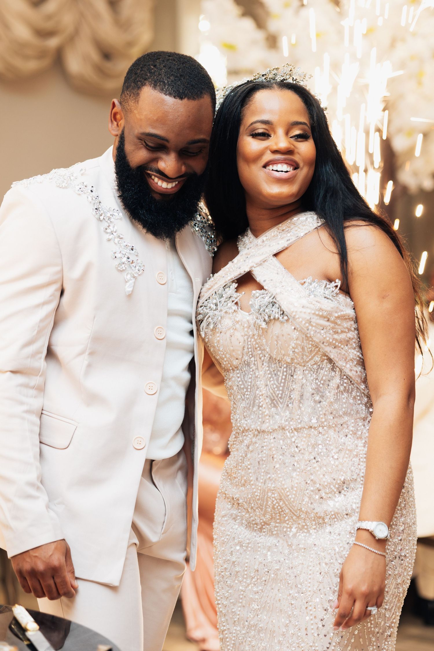 Elegant couple in sparkling wedding attire smiling joyfully, bride wearing crystal gown and tiara, groom in white jeweled suit.