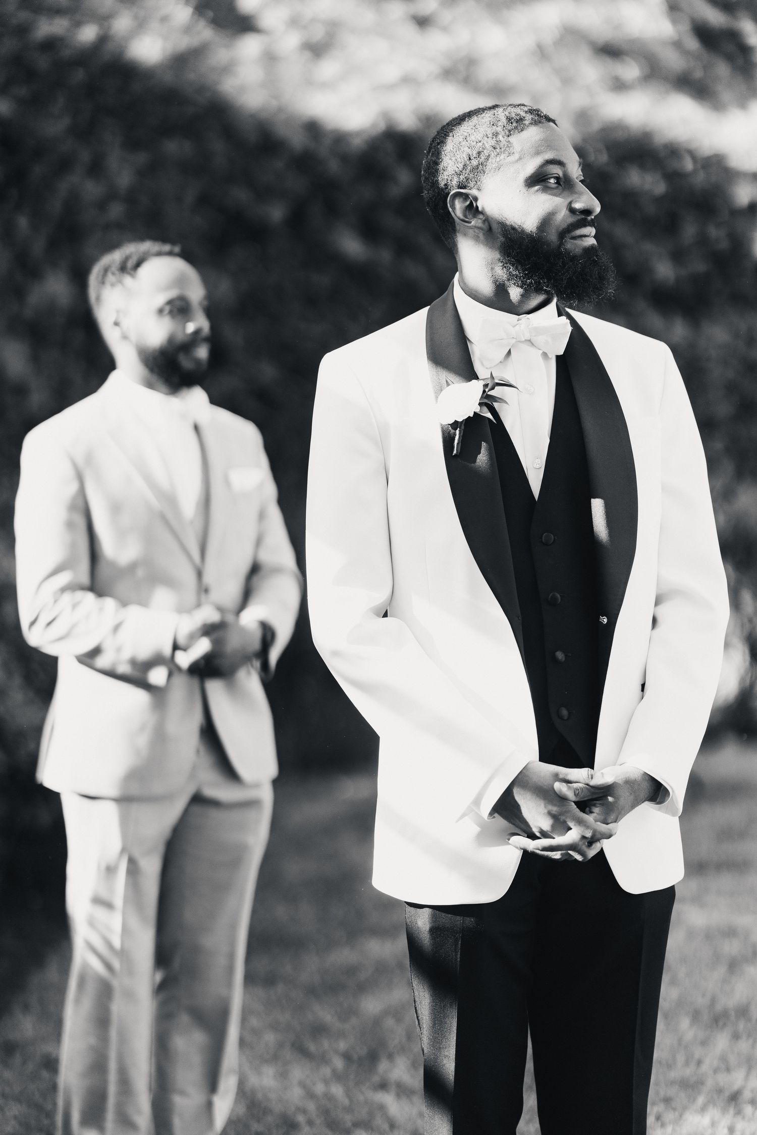 Two grooms in elegant suits stand outdoors in a black and white wedding photo.